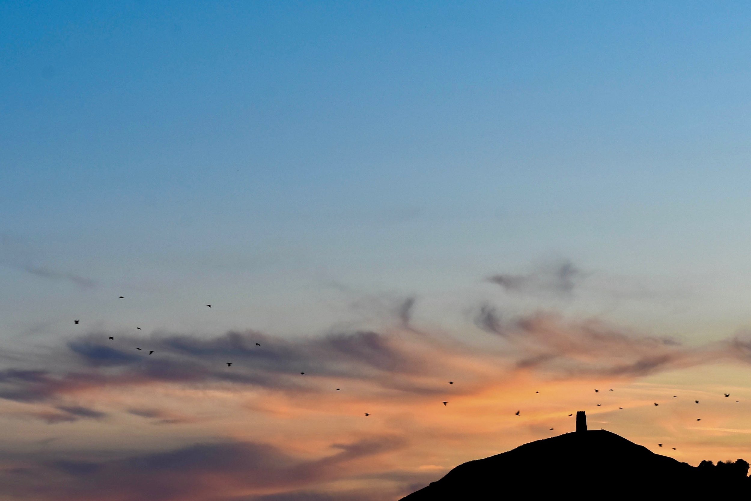 The Glastonbury Tor, an important spiritual site in Christianity, Celtic mythology, and new-age Goddess Worship, is silhouetted against the vibrant evening sky.