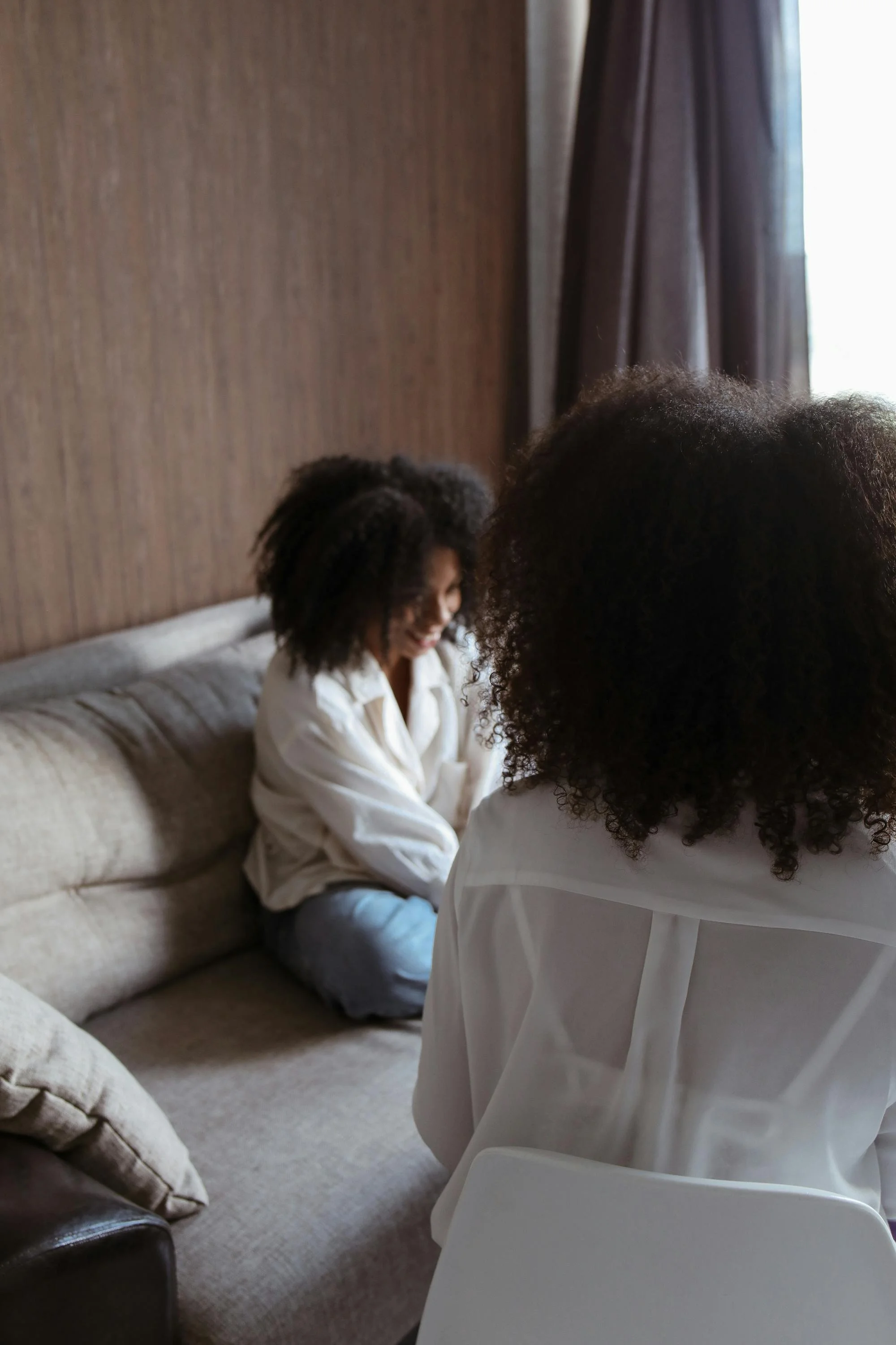 Two women with curly hair sitting on a beige couch, smiling and talking indoors near a window with curtains.
