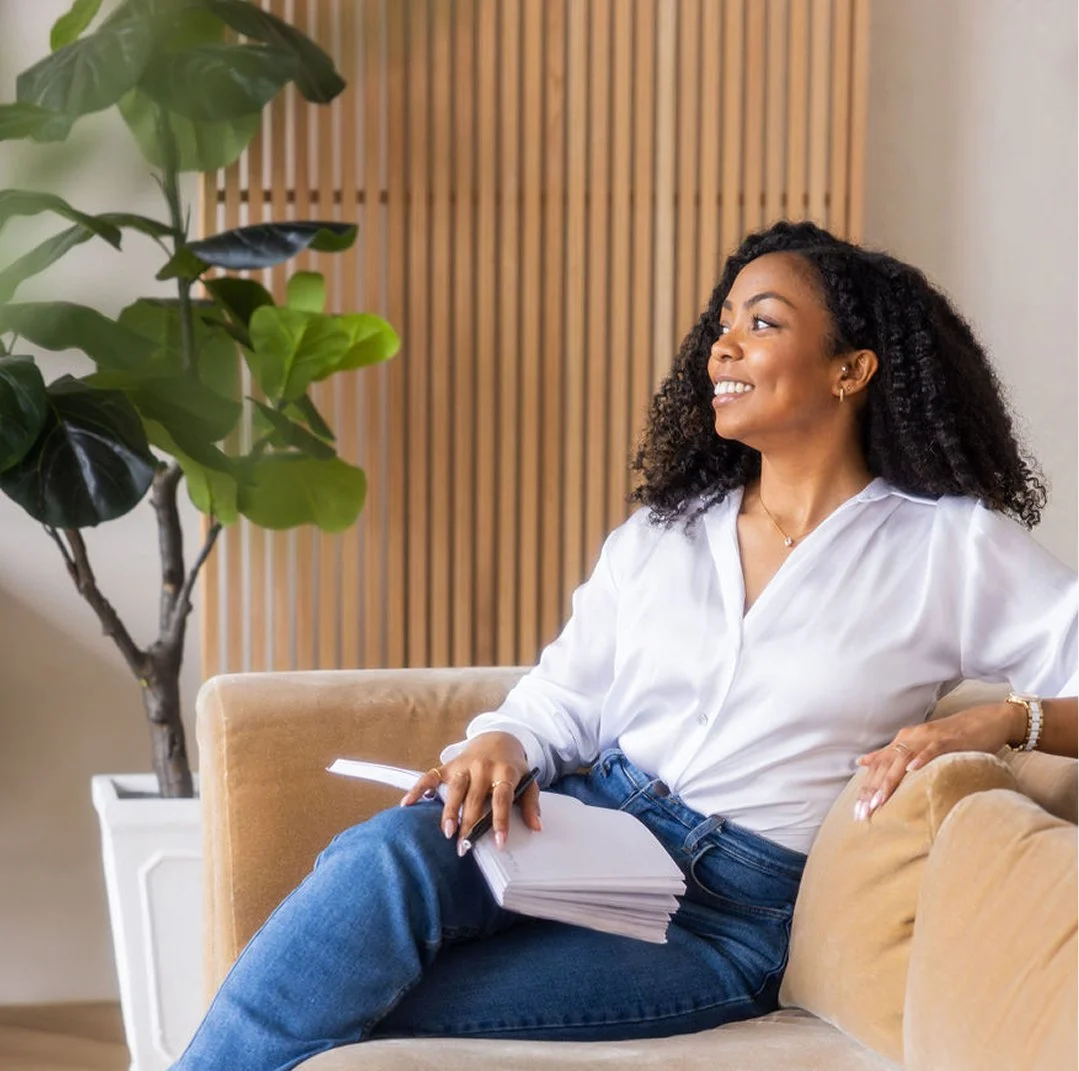 A woman with curly hair sitting on a beige couch, holding a notebook, smiling, in a room with a potted plant and wooden wall paneling.