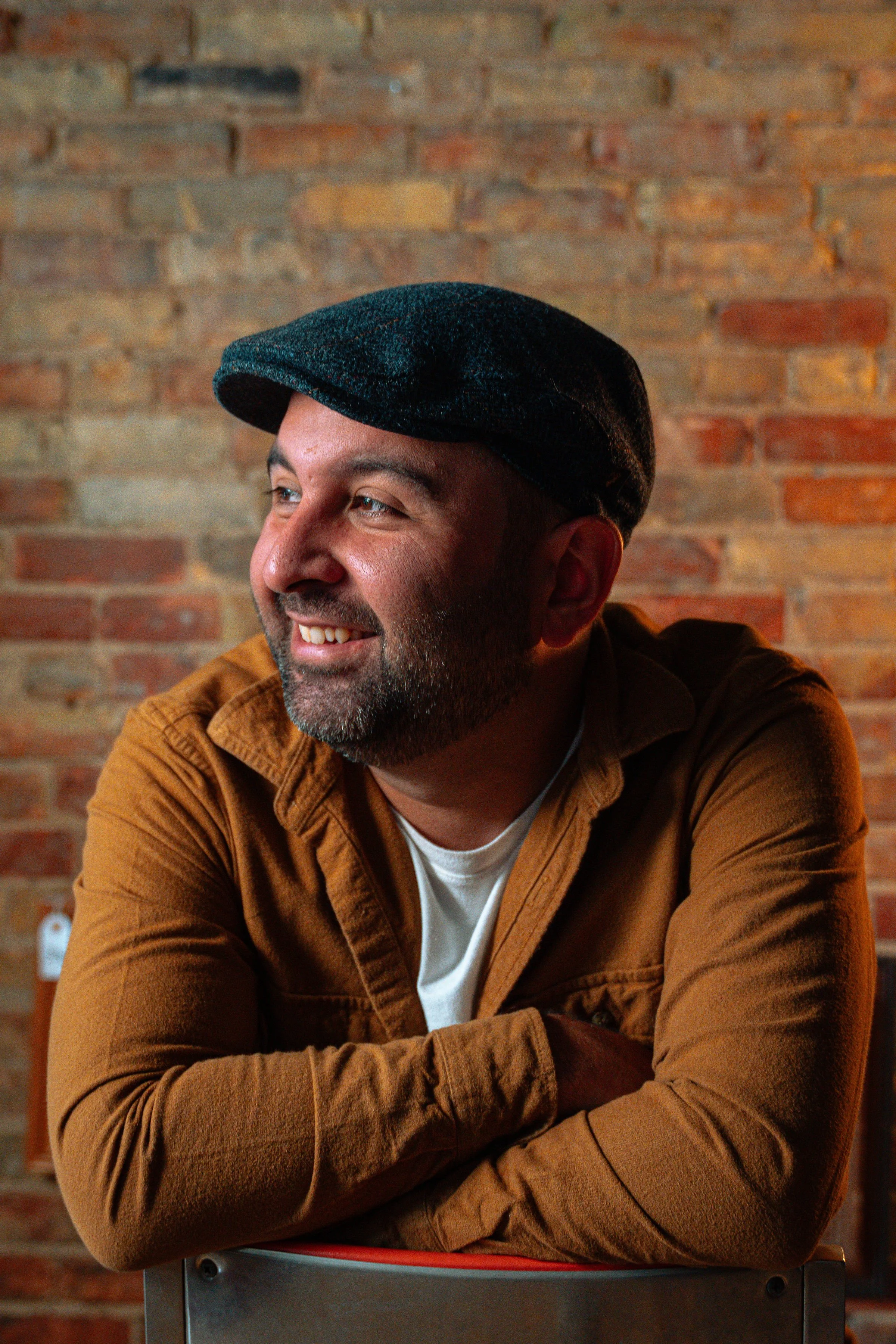 A smiling man with a beard wearing a black flat cap and a brown jacket, sitting with his arms crossed, in front of a brick wall.