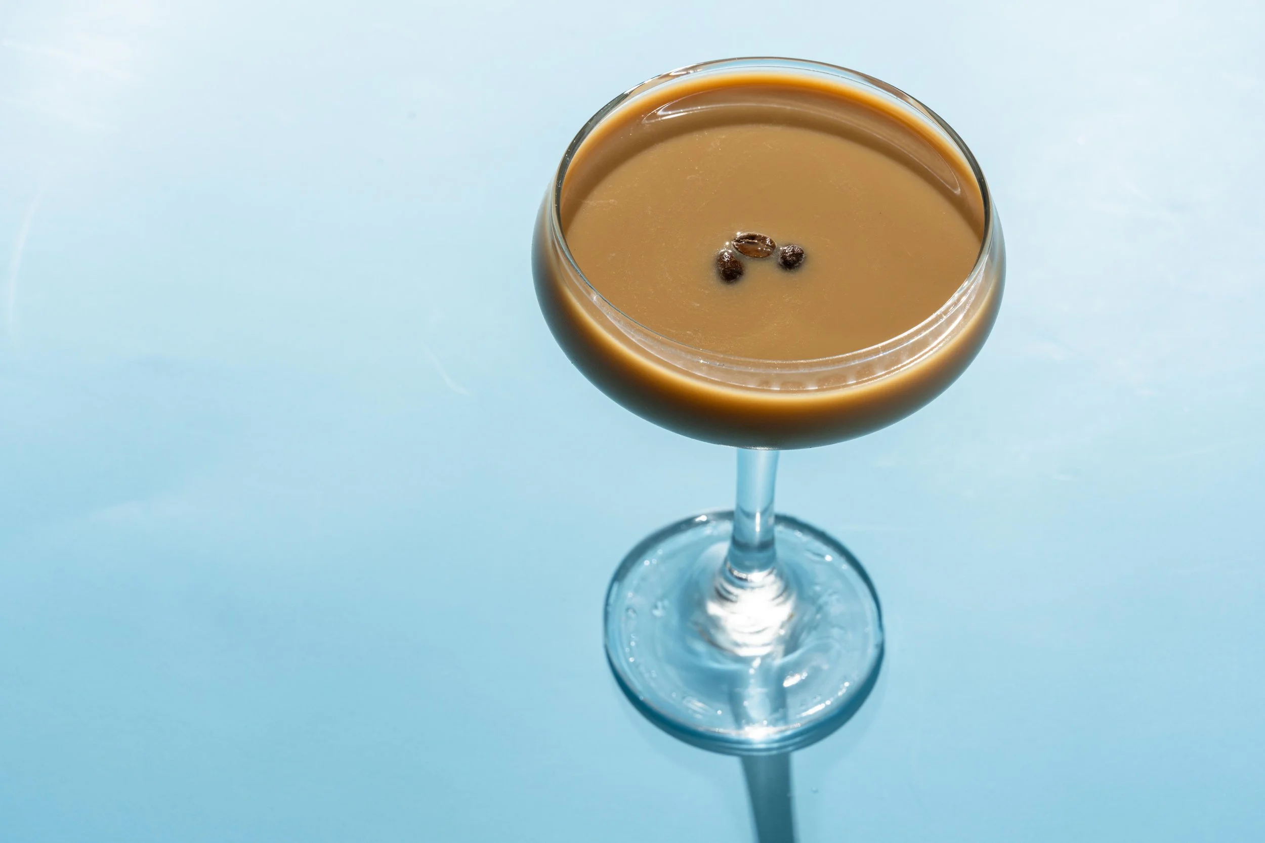 A cocktail glass filled with a light brown beverage, garnished with coffee beans floating on top, placed on a light blue surface.