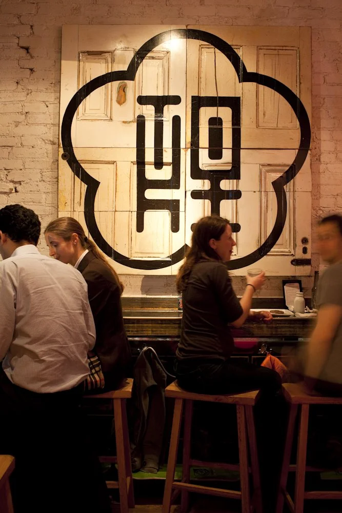 People sitting on bar stools and socializing in a dimly lit bar with exposed brick walls. A large mural with a stylized design is hanging on the wall behind them.