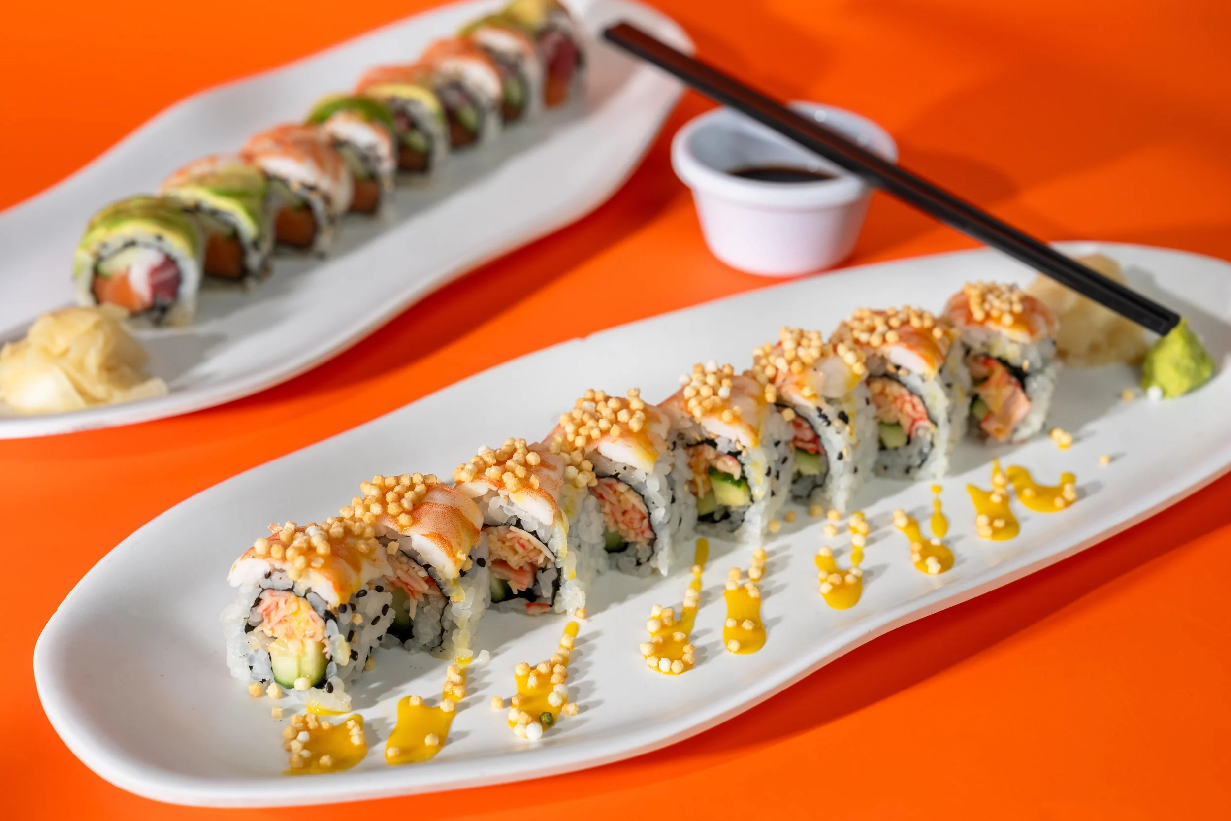 Close-up of two white rectangular plates filled with sushi rolls on an orange table, with a small white dish of soy sauce and black chopsticks resting on one plate.