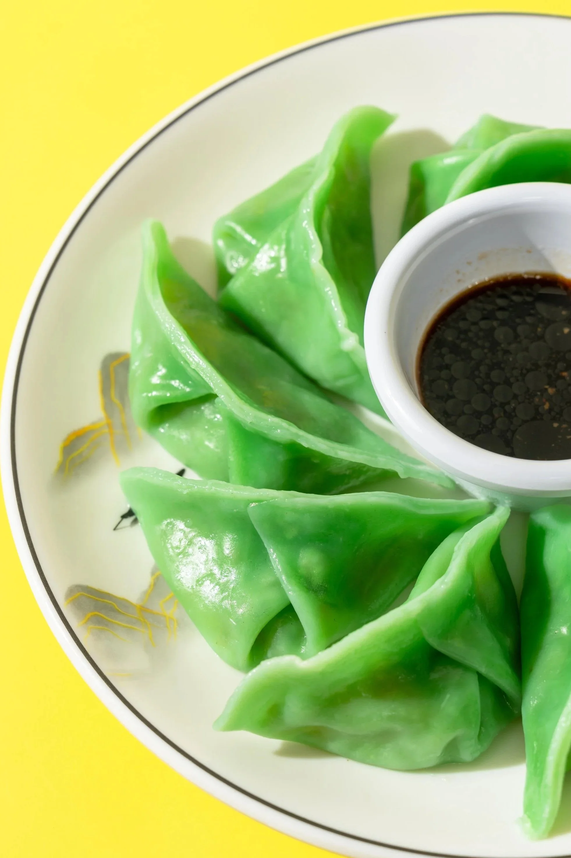 Bright green steamed dumplings on a white plate with a small bowl of soy sauce on the side, against a yellow background.