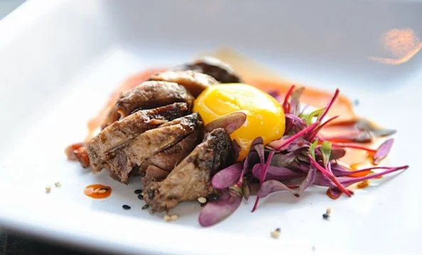 A dish with sliced cooked meat, a raw egg yolk on top, and a small side salad with purple microgreens.