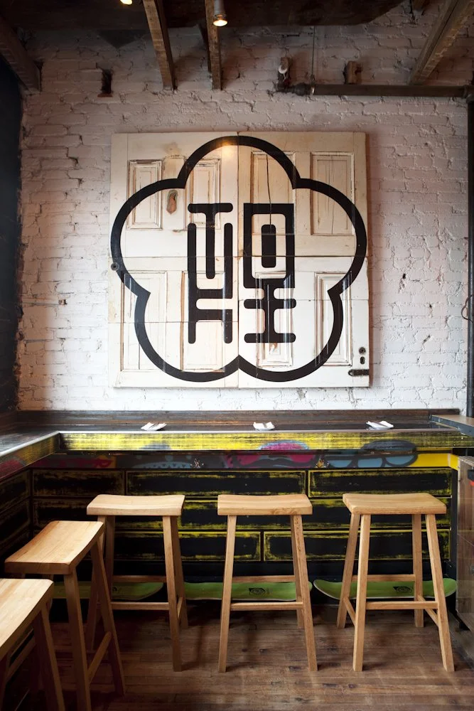 Interior of a bar with a white brick wall featuring a large black graphic of a flower with stylized characters inside, three wooden bar stools in front, and a graffiti-style artwork on the front of the bar counter.