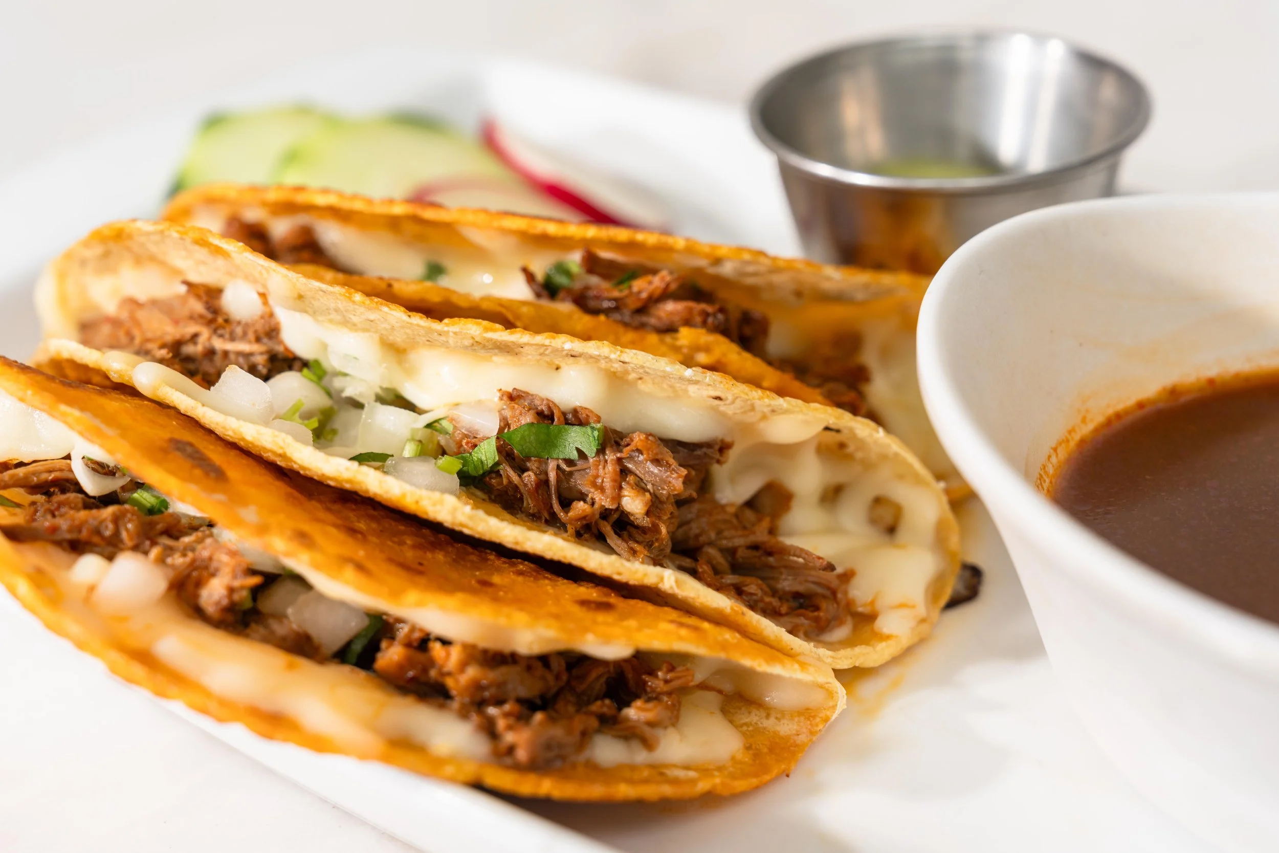 Close-up of two beef tacos with melted cheese, chopped onions, and green herbs, served with a side of dark dipping sauce in a small white bowl and a metal cup of lime juice.