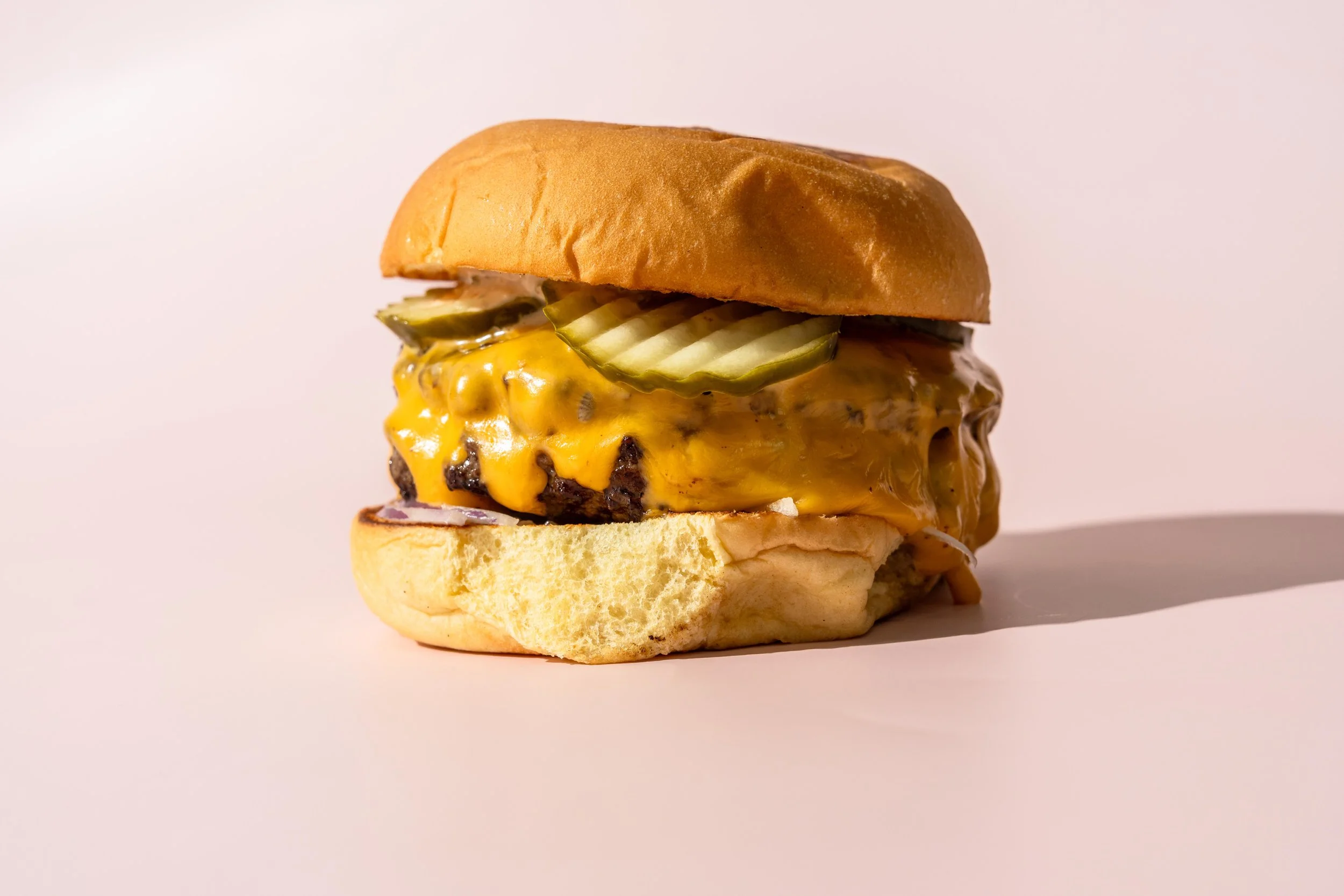 A close-up image of a cheeseburger with pickles, melted cheese, bacon, and a bun, on a plain white background.