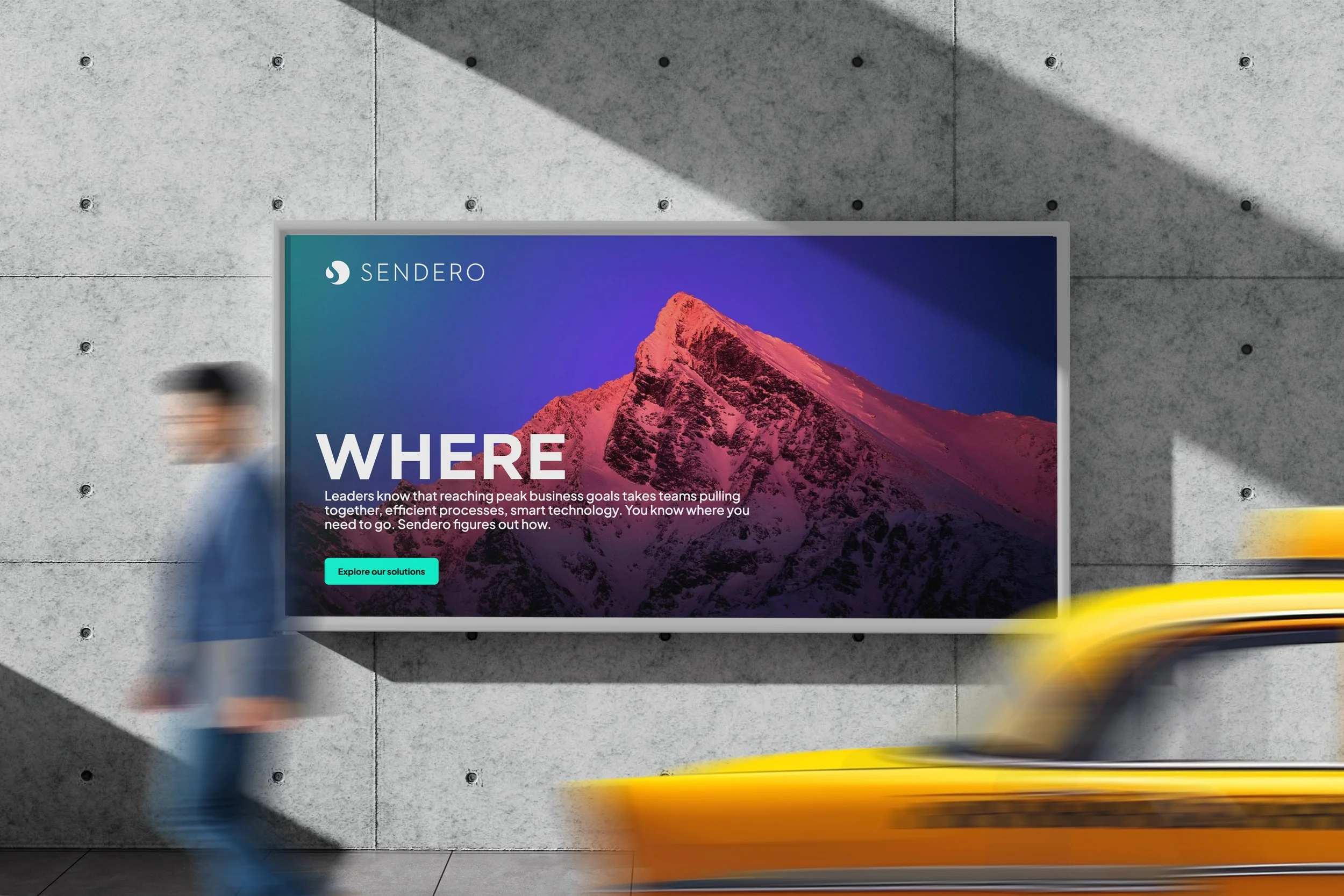 A digital billboard displaying an advertisement for Sendero, featuring a mountain peak with a pinkish hue against a dark blue sky. There are blurred figures of people walking by in front of the billboard, on a concrete wall background.