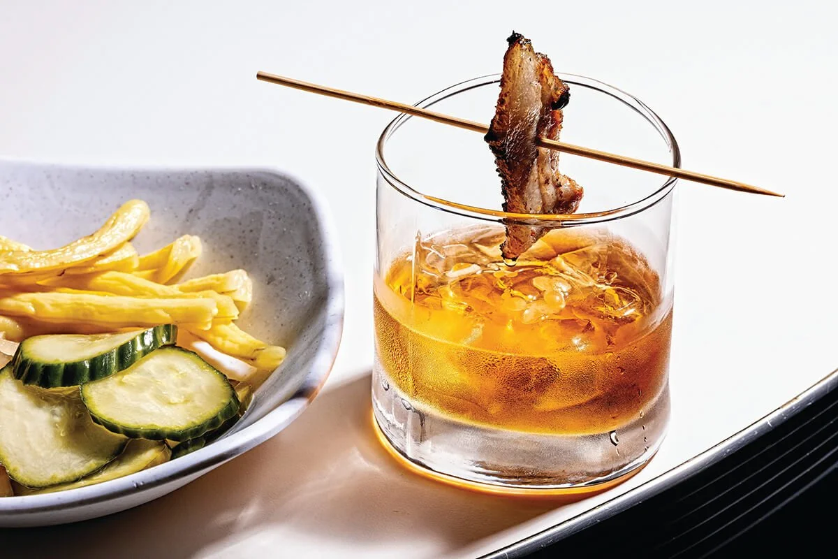 A glass of whiskey with ice and a strip of bacon on a skewer, served on a white tray. To the side, a bowl containing sliced cucumbers and yellow pickled vegetables.