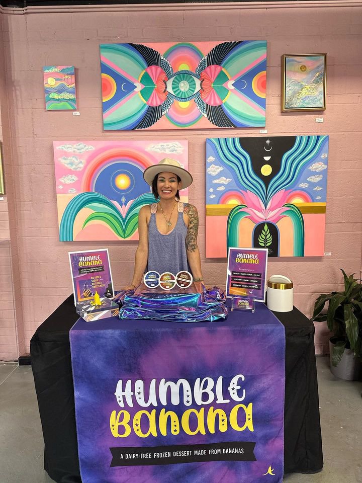 A woman standing behind a table that displays a banner for 'Humble Banana,' a dairy-free frozen dessert made from bananas. The woman is smiling, wearing a hat and a tank top, with colorful abstract paintings hanging on a pink wall behind her.