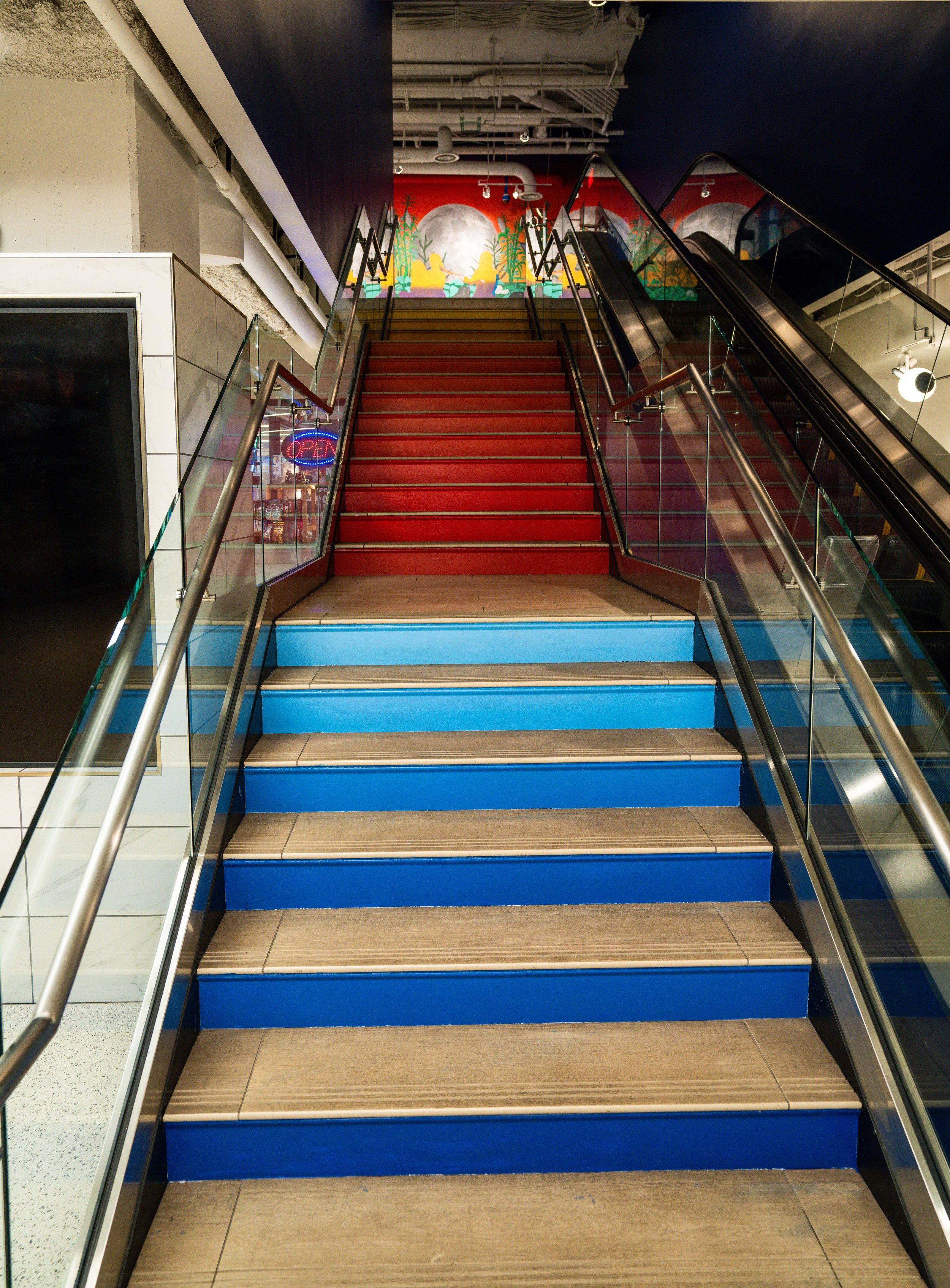 An escalator with red steps and blue trim at the bottom, leading up to a colorful mural with abstract trees, a moon, and sky, in an indoor shopping mall or entertainment complex.