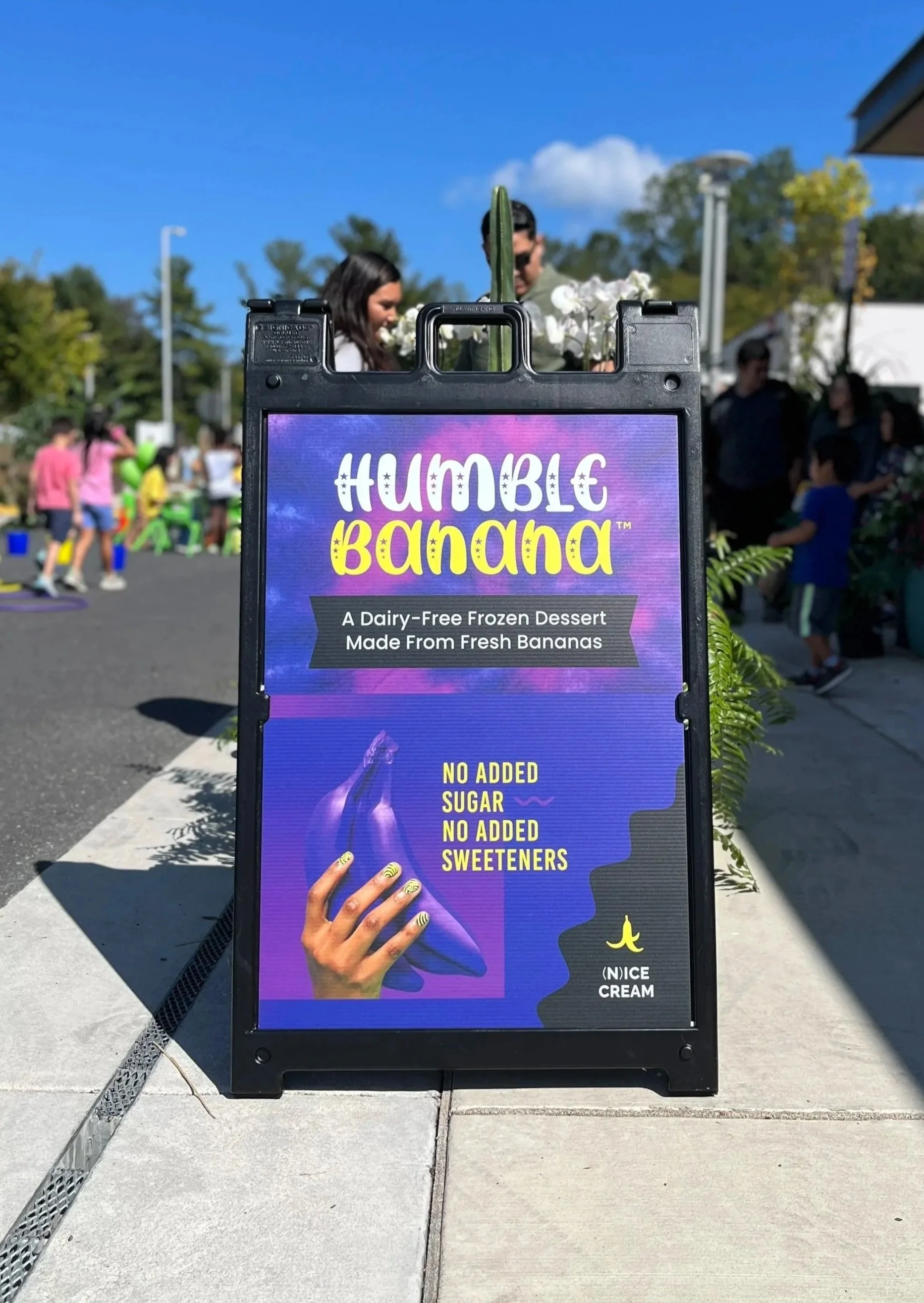 A promotional sign for 'Humble Banana' frozen dessert made from fresh bananas, emphasizing no added sugar or sweeteners, with a colorful purple and blue background and a stylized hand pointing to the text.