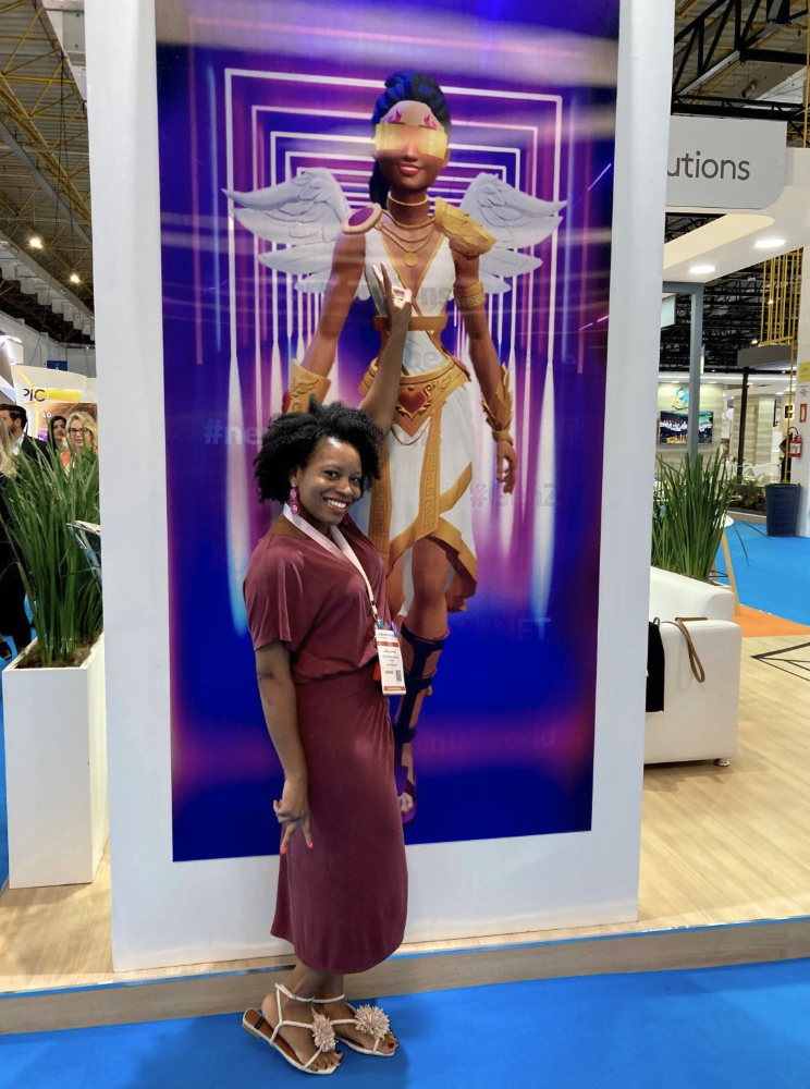 A woman in a burgundy dress and beige sandals standing in front of a large digital display of a fantasy character with wings, a gold and white outfit, and a futuristic background.