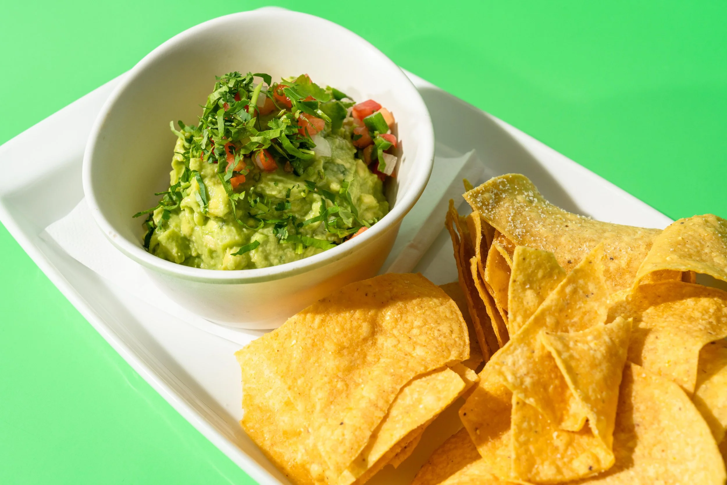 Bowl of guacamole garnished with chopped tomatoes, onions, and cilantro, next to a side of tortilla chips on a white tray, set against a green background.