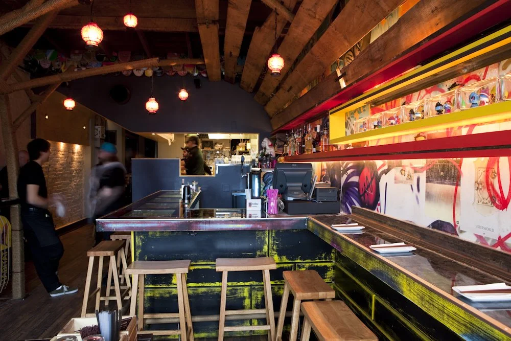 Interior of a restaurant or bar with a wooden ceiling, hanging red lanterns, a counter with chairs, and a colorful graffitied wall.