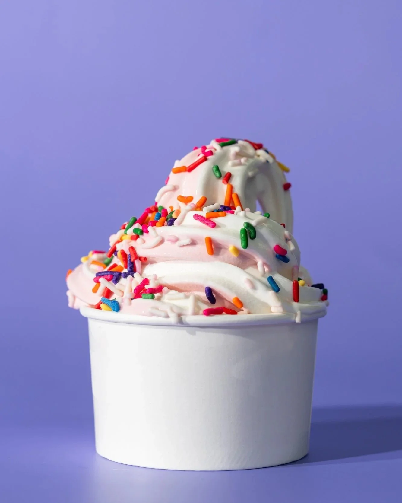 Cup of pink and white ice cream with colorful sprinkles on top against a purple background.