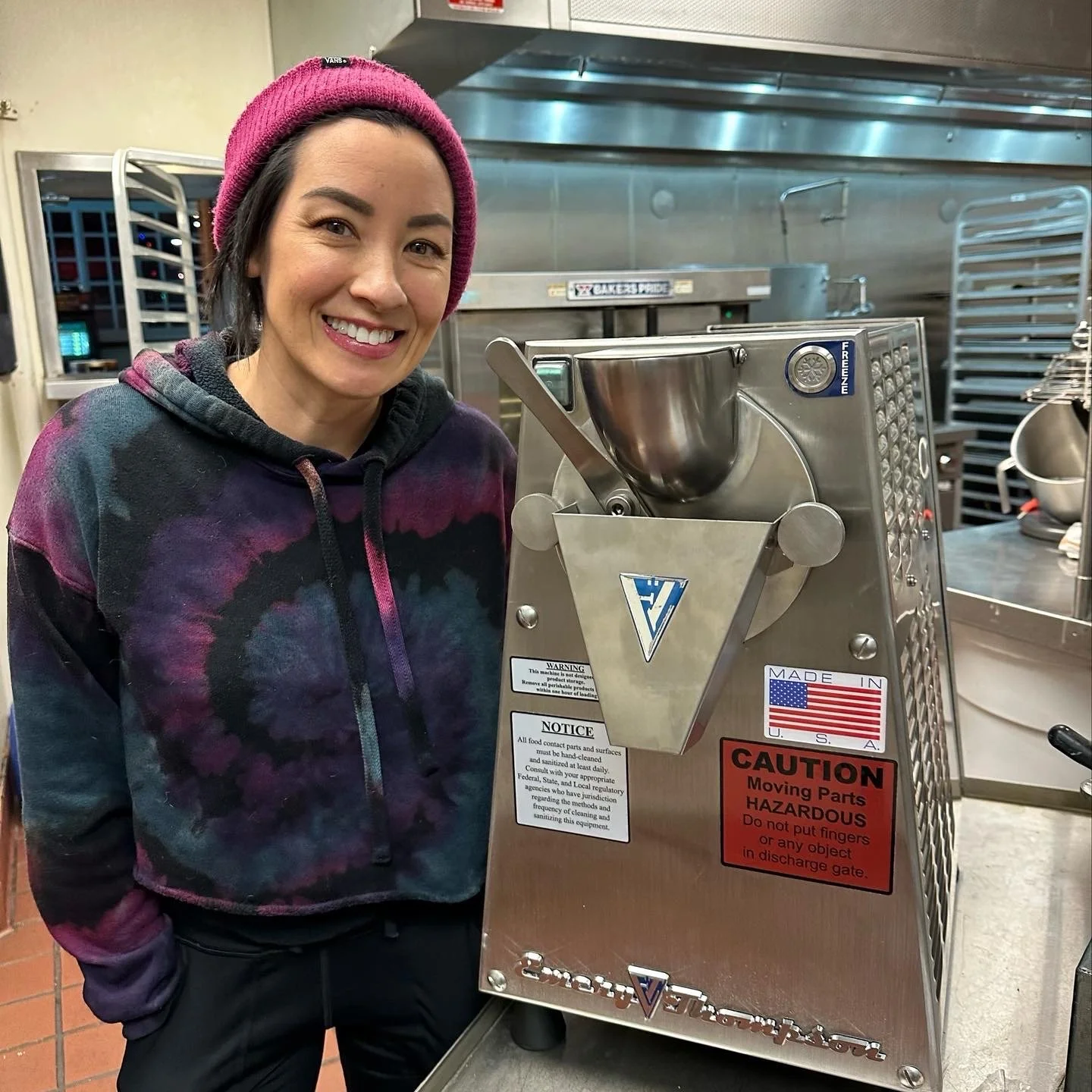 A woman with a pink beanie and a tie-dye hoodie smiling in a kitchen or commercial setting, standing next to a large industrial machine with warning labels.