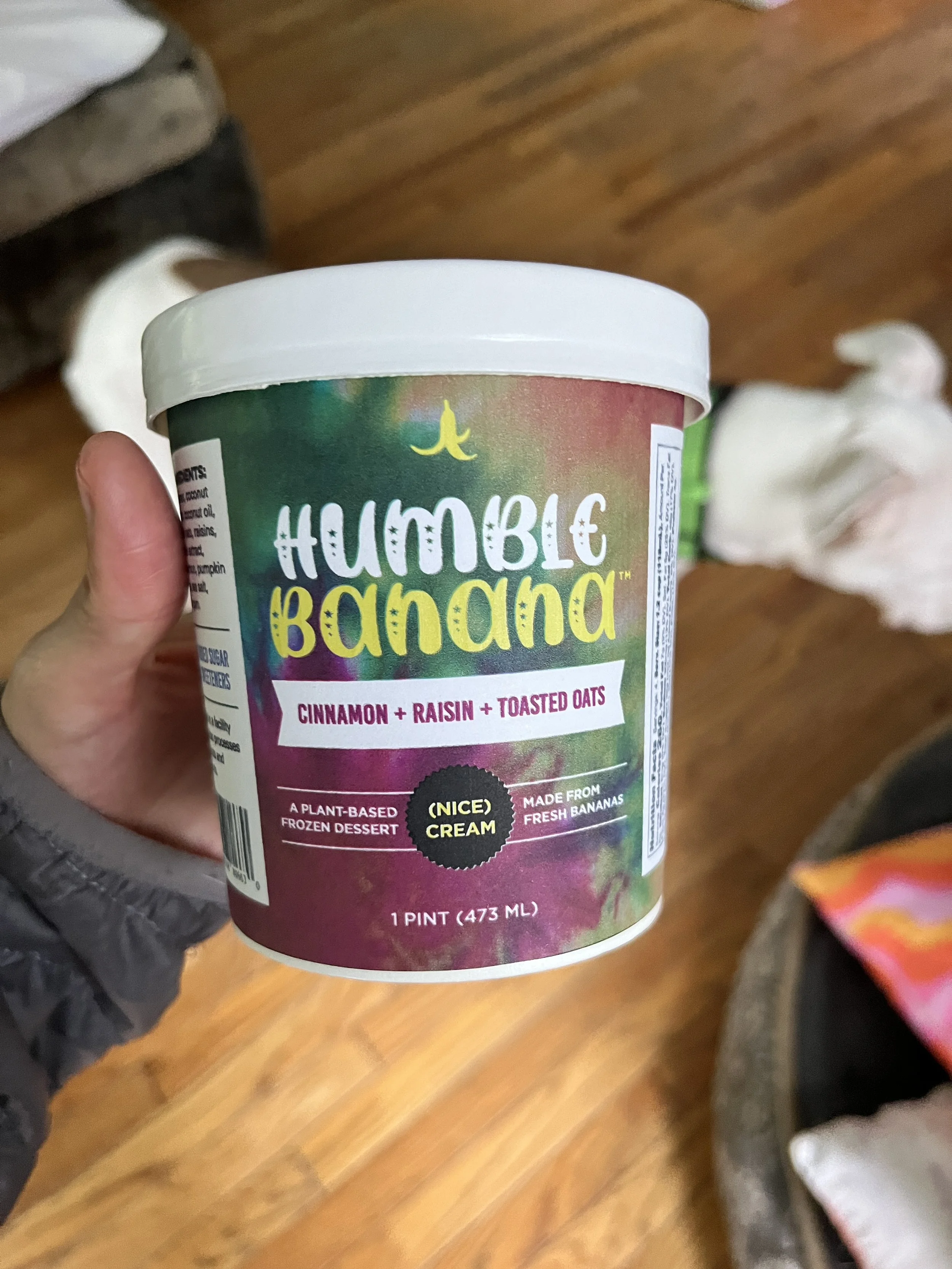 A hand holding a pint container of 'Humble Banana' frozen dessert with cinnamon, raisin, and toasted oats, on a wooden floor.