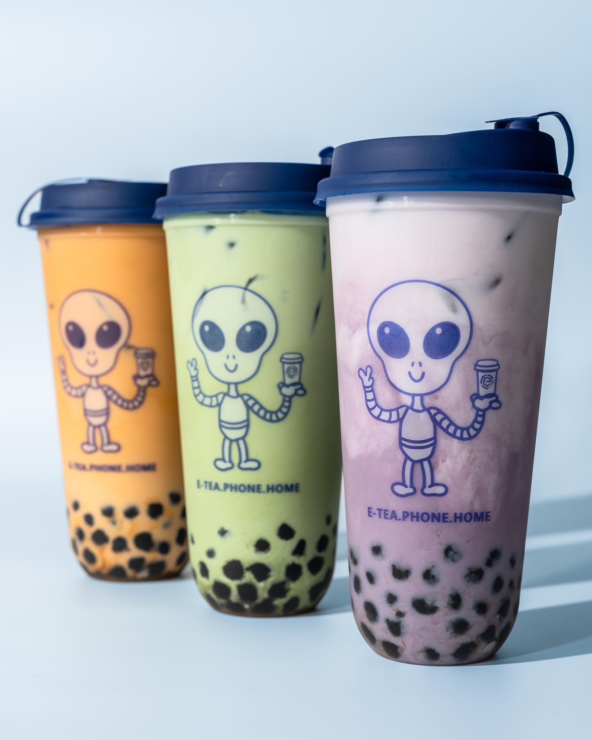 Three cups of bubble tea with tapioca pearls, featuring cartoon alien face logos, in yellow, green, and pink colors, on a light blue background.