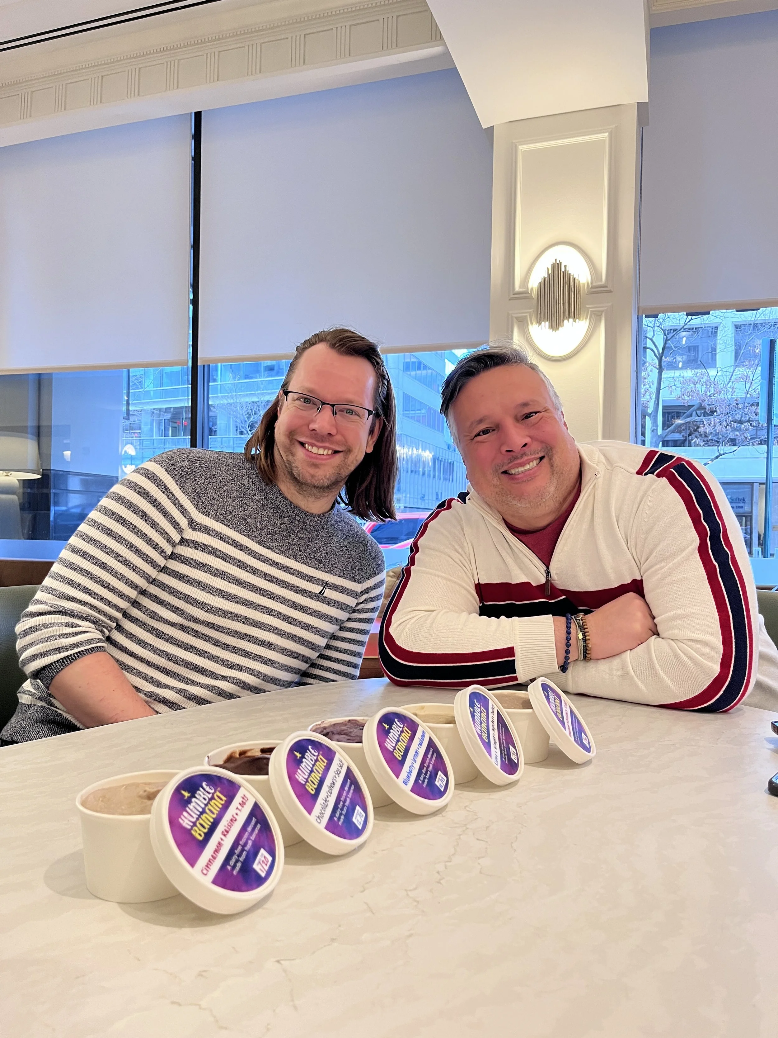Two smiling men sitting at a table in a cafe, with several tubs of ice cream in front of them. The background shows large windows with a city view and modern decor.