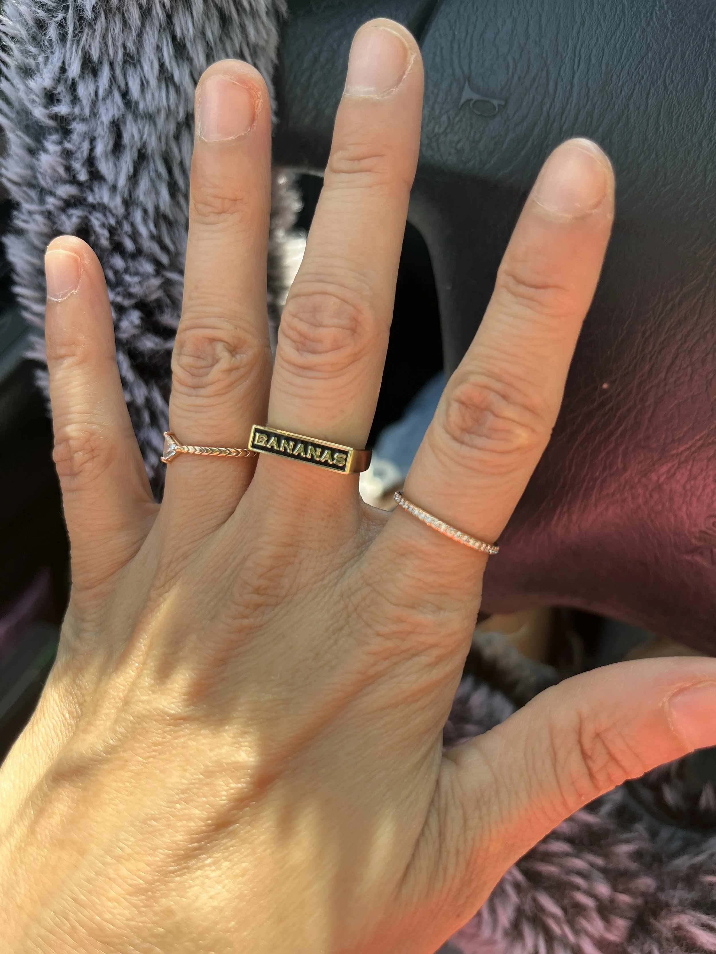 Close-up of a person's hand with three rings, one reading 'BANANAS,' and their nails are trimmed but slightly dry or peeling, resting on a black steering wheel in a vehicle, with a fuzzy gray object in the background.