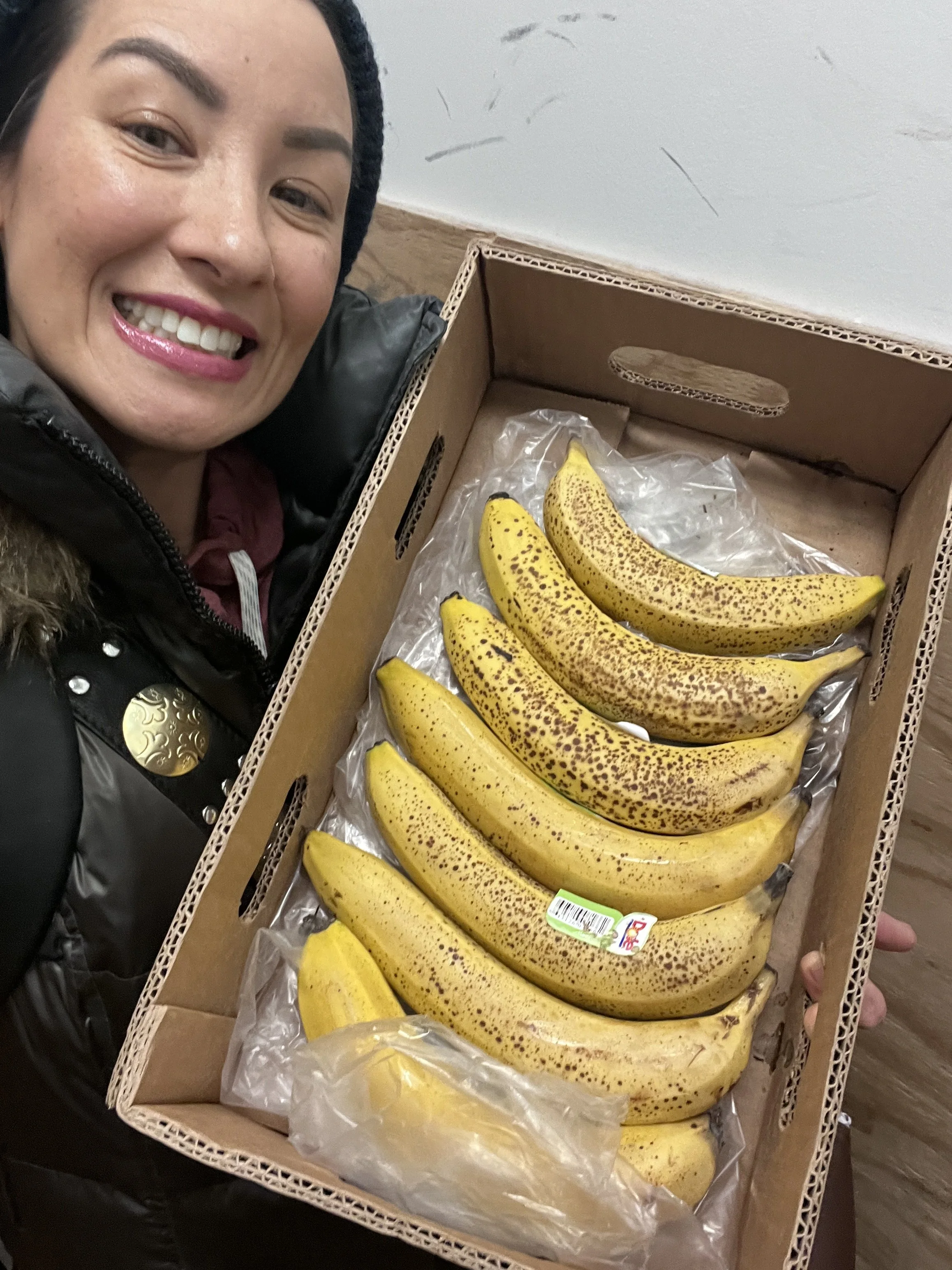 A woman smiling for the camera while holding a cardboard box filled with yellow bananas with brown spots on a plastic lining.