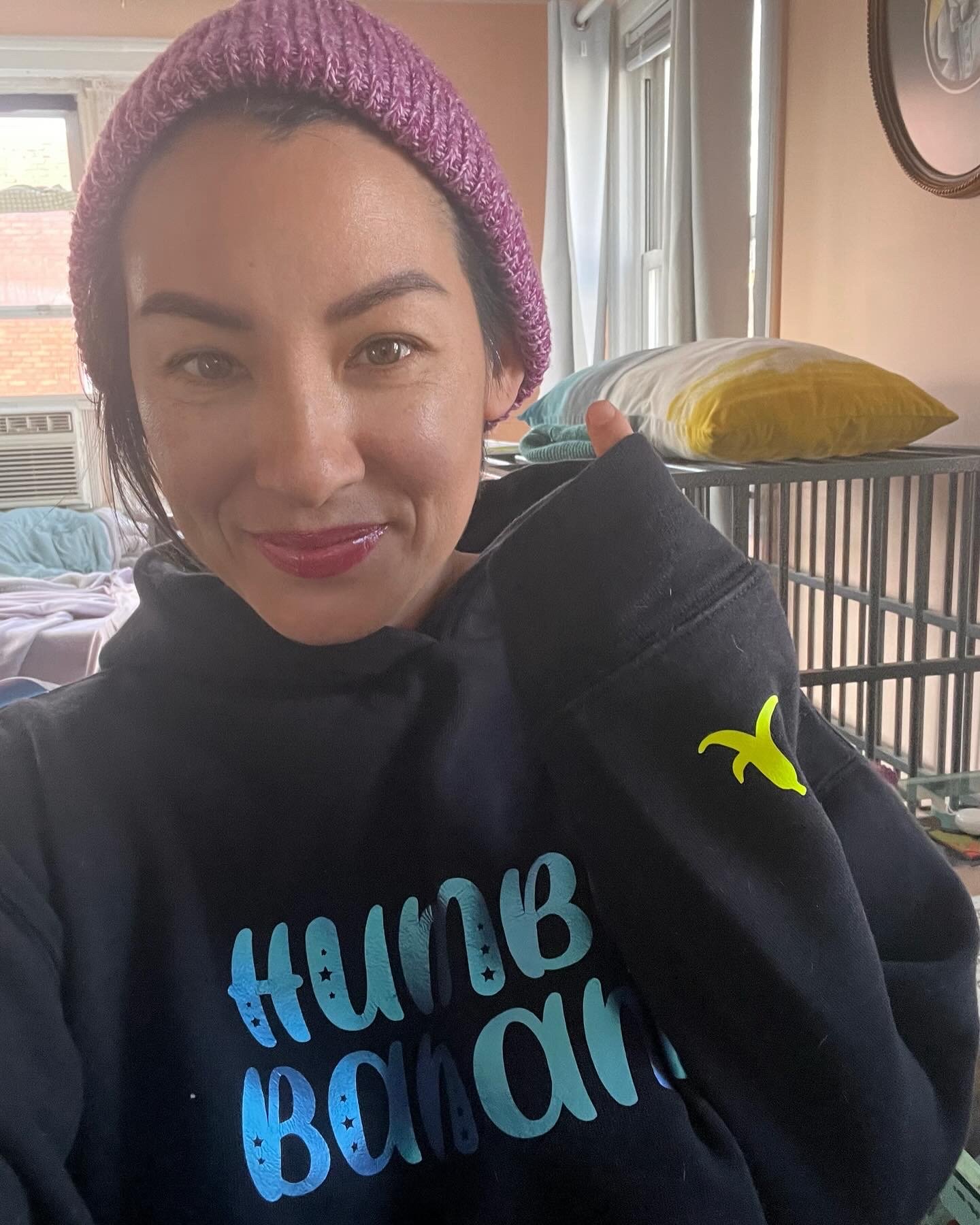 A woman wearing a pink knit hat and a black hoodie with blue lettering, smiling indoors near a bed and large windows, with pinkish walls and sunlight through the curtains.