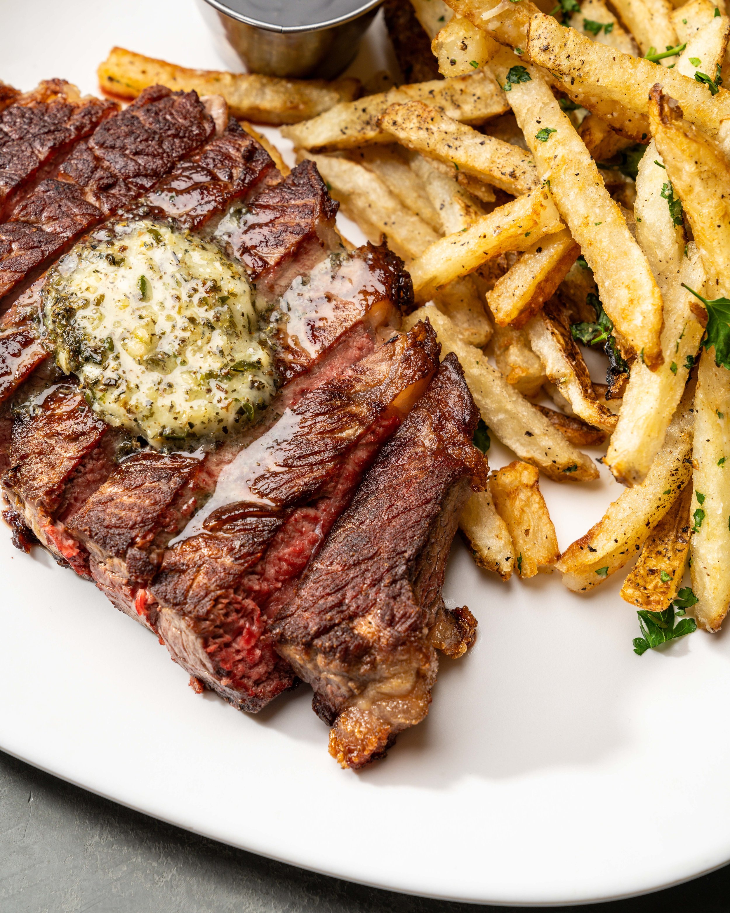 A plate of grilled steak topped with herb butter, served with seasoned French fries garnished with parsley, and a small metal cup of dipping sauce.