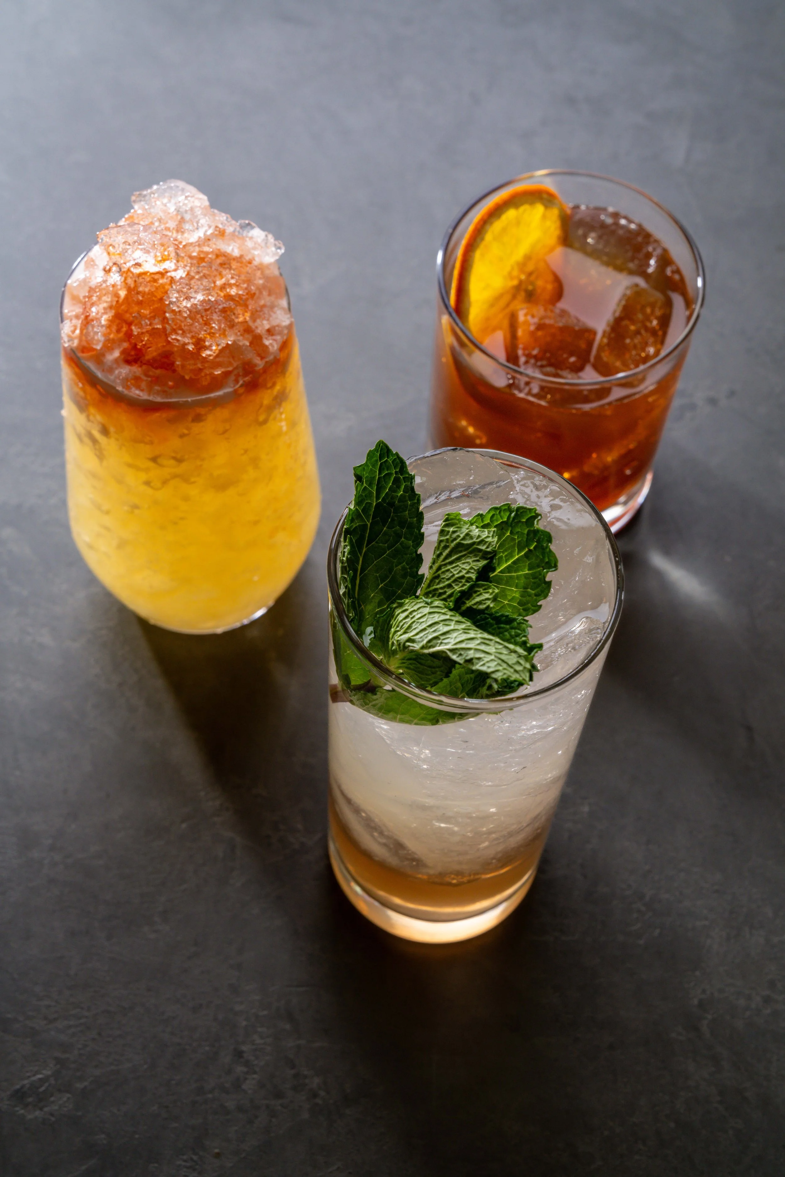 Three cocktails on a dark surface. The left contains a yellow drink garnished with crushed ice on top, the middle has a dark amber drink with ice and an orange slice, and the right shows a clear drink with ice, garnished with a sprig of mint.