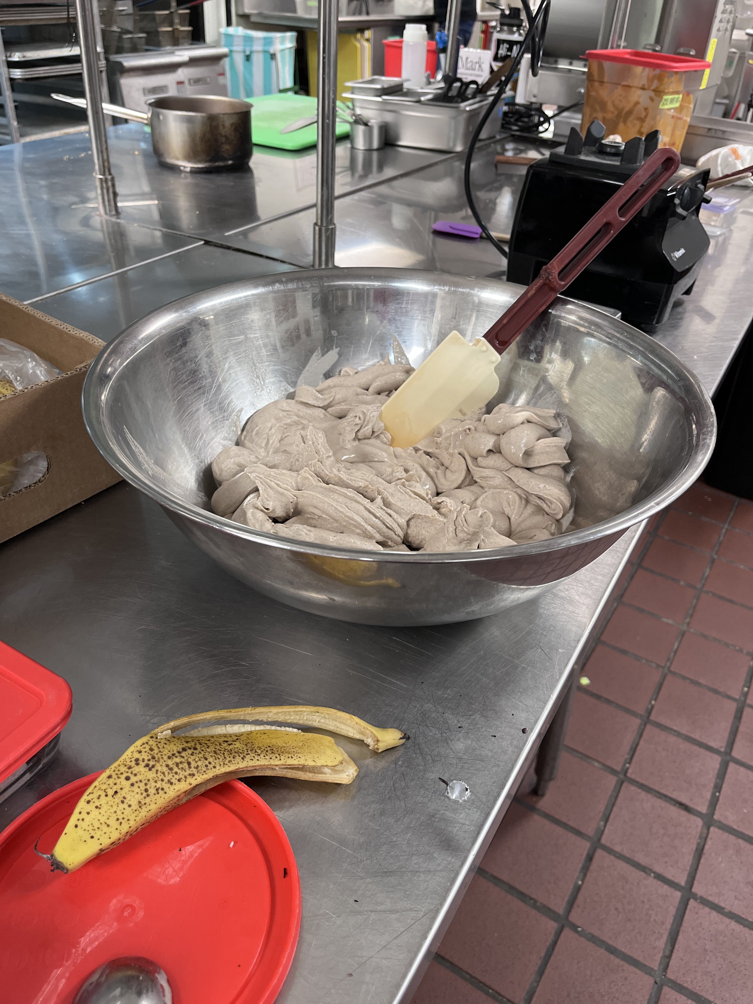 A stainless steel mixing bowl with a yellow spatula inside, containing a light brown, creamy mixture, on a stainless steel countertop in a commercial kitchen. There are bananas and a red plastic container nearby, and various kitchen equipment in the background.