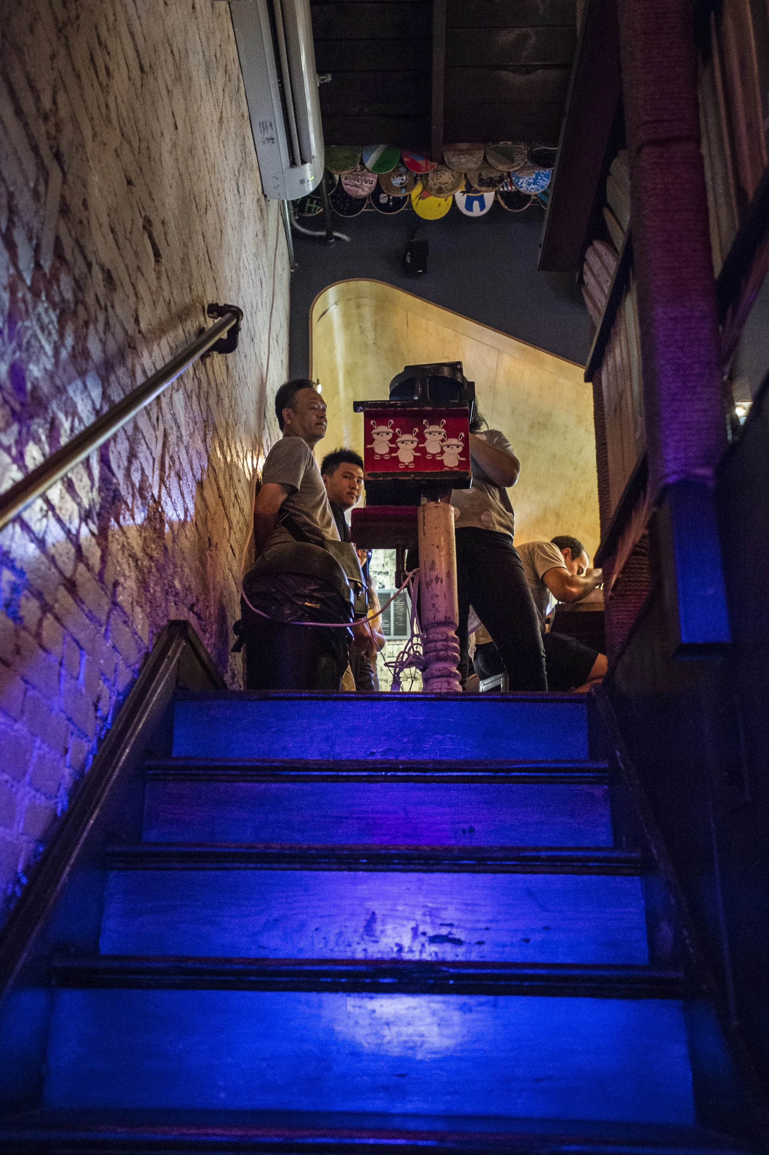 People standing on a staircase inside a dimly lit venue, with a brick wall on one side and decorative hats hanging on the ceiling.