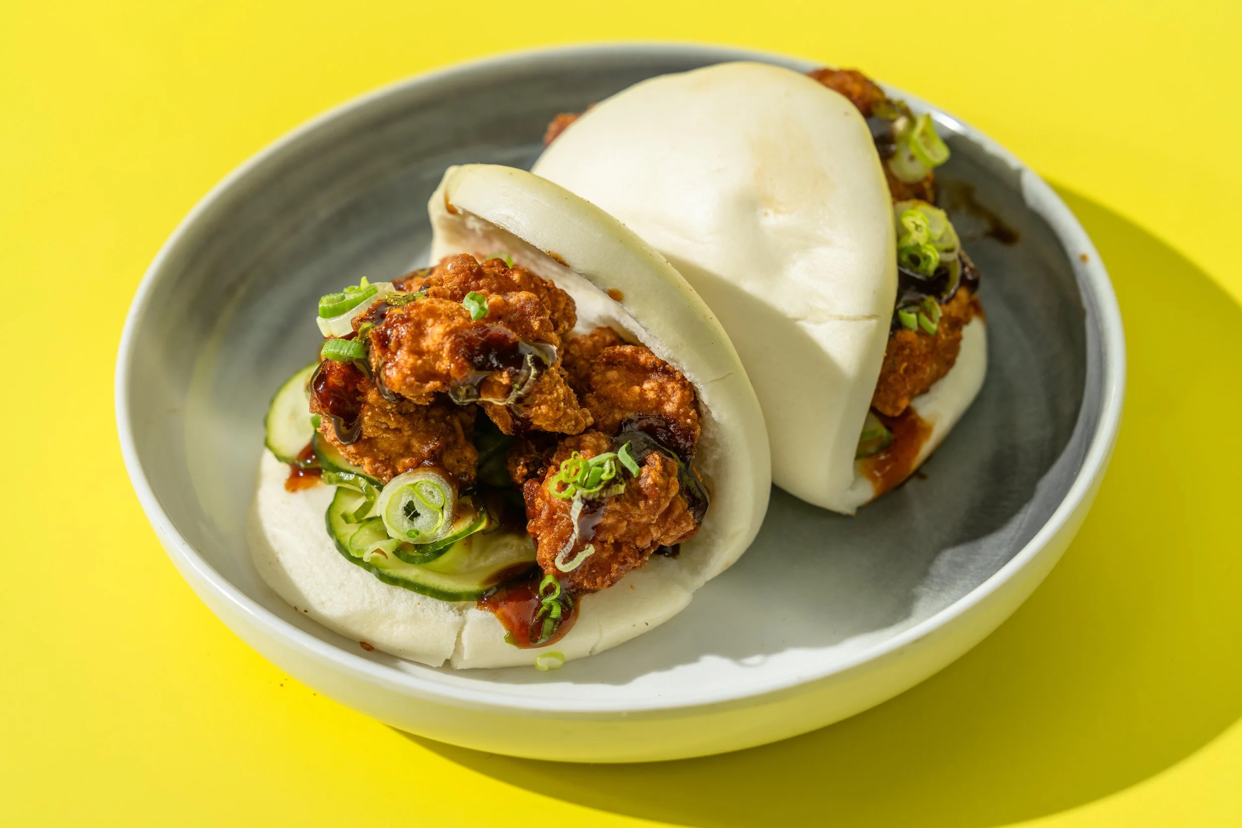 Chinese steamed buns filled with fried chicken, sliced green onions, and drizzled with a dark sauce, served in a white bowl on a yellow background.