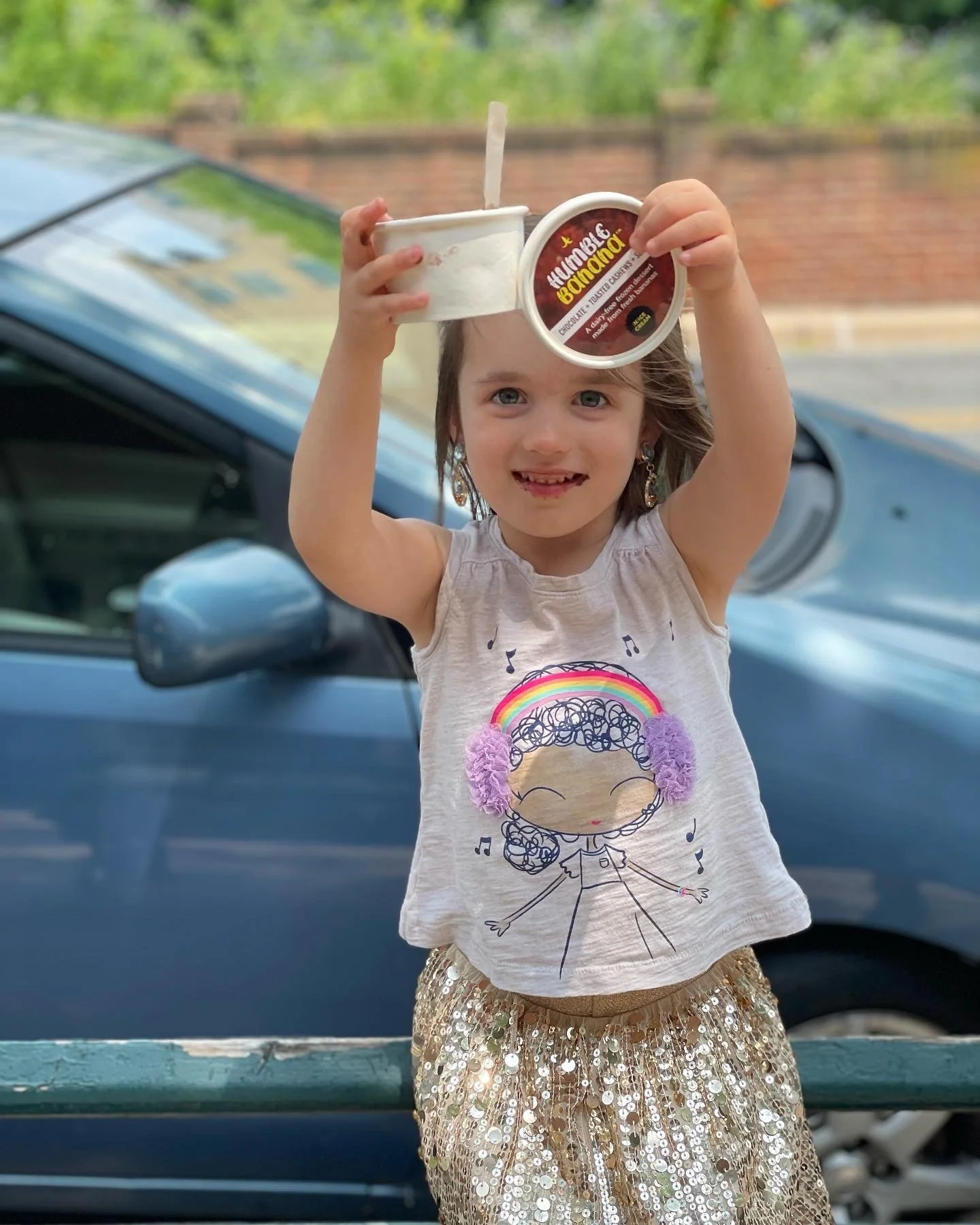 A young girl with blue eyes, wearing a white shirt with a cartoon girl and musical notes and a gold sequin skirt, holds up two containers of chocolate banana ice cream outdoors near a blue car, with a blurred background of greenery and a brick wall.