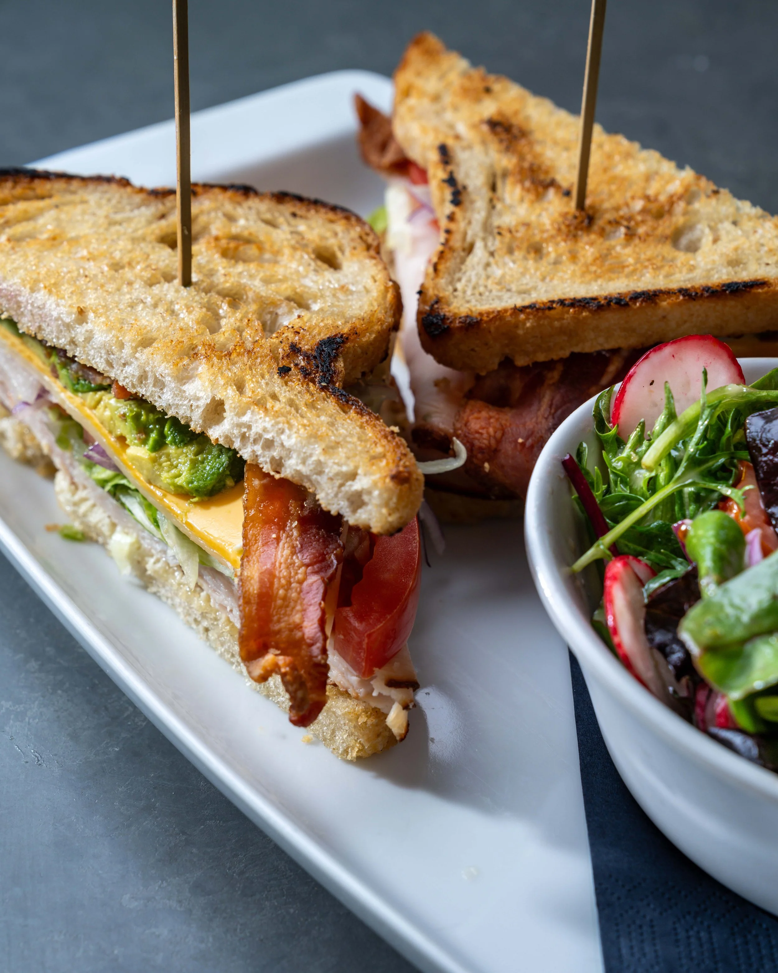 Close-up of a sandwich on a white rectangular plate, with a side salad in a small white bowl, all on a dark gray surface.