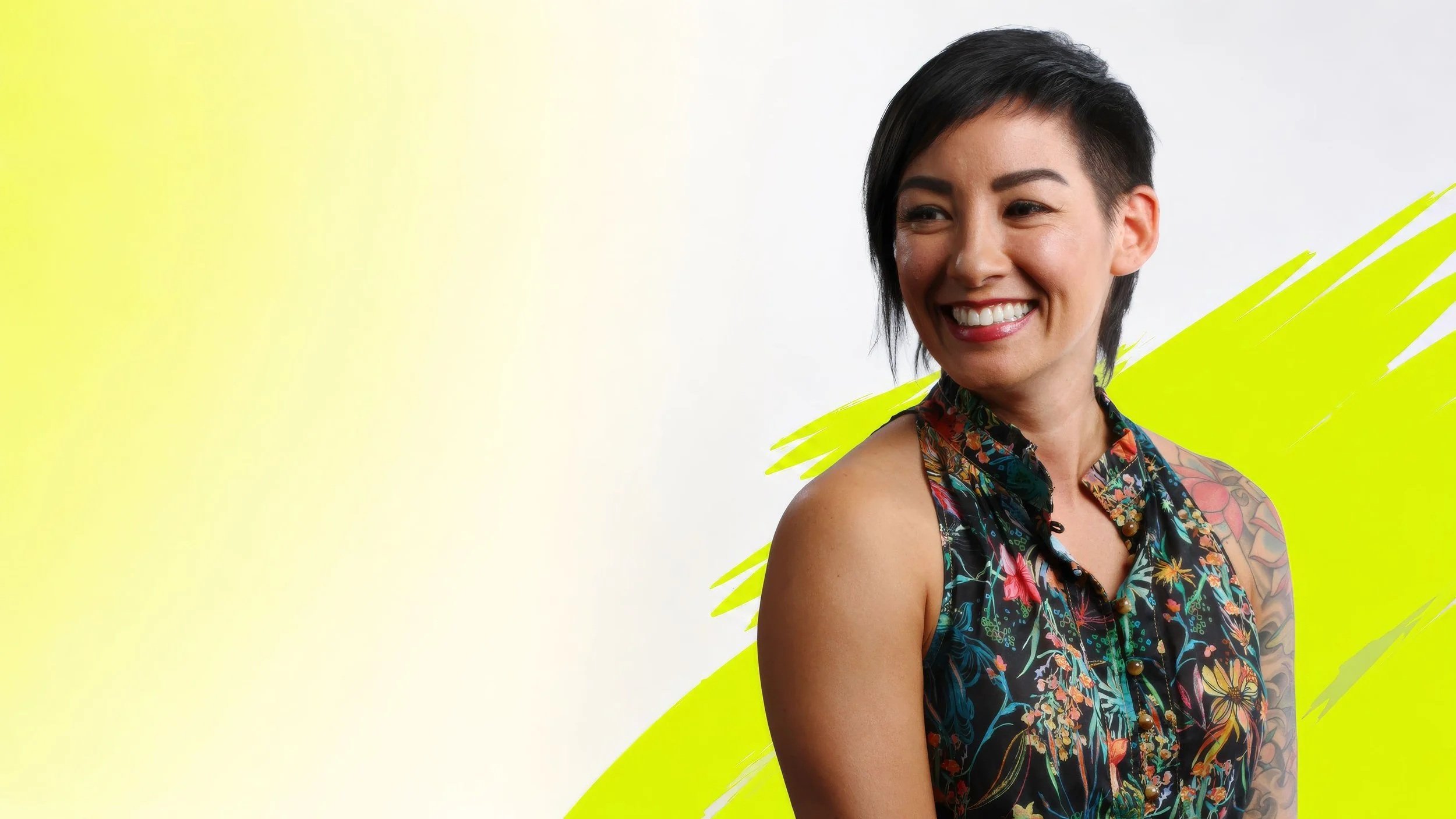 Smiling woman with short dark hair, wearing a floral sleeveless top, standing against a yellow and white background with yellow paint strokes.