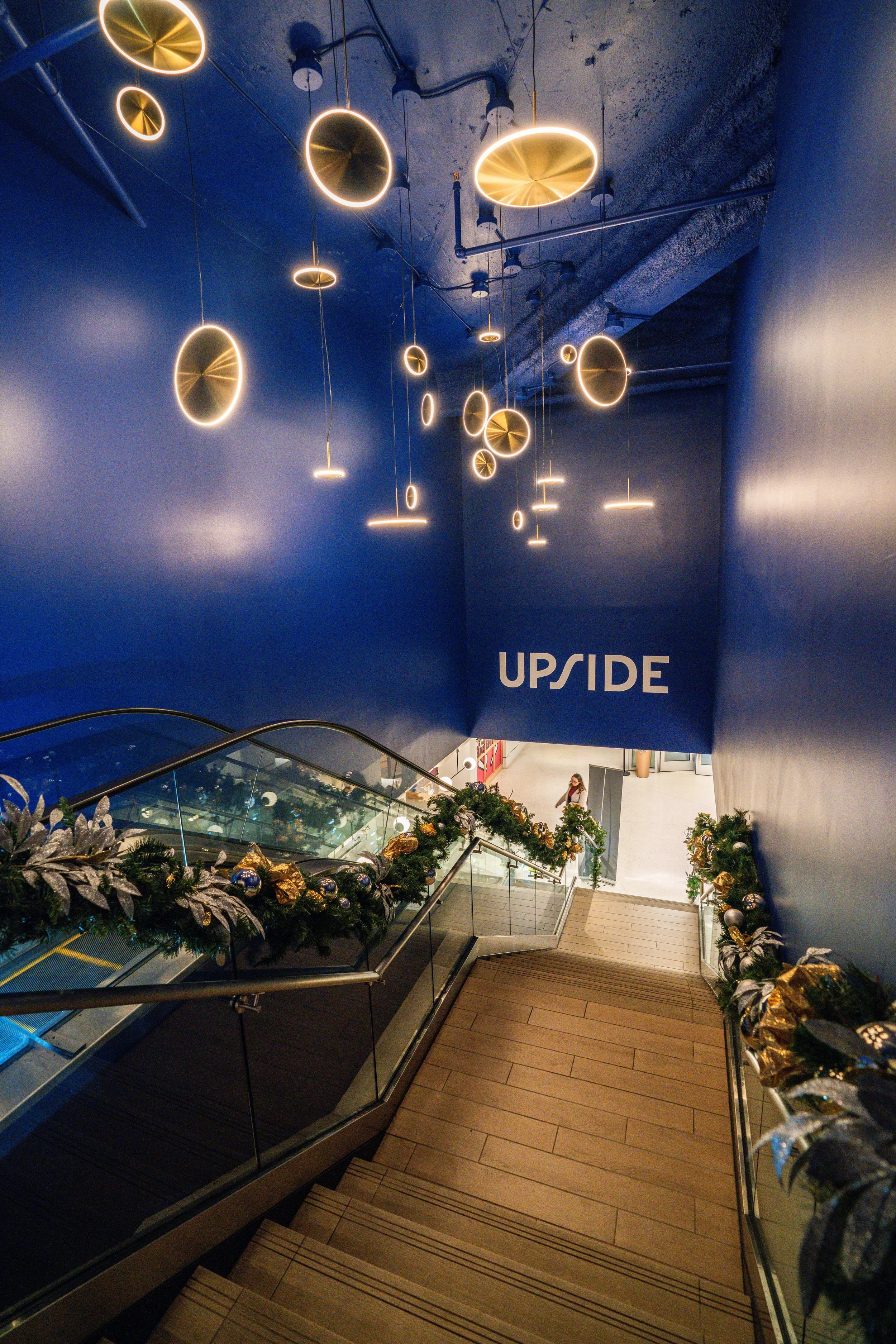 Decorative golden and silver ornaments along a stairway railing with holiday greenery, leading to an upper level with modern hanging circular and oval light fixtures and a blue wall with the word 'UPSIDE' in white.