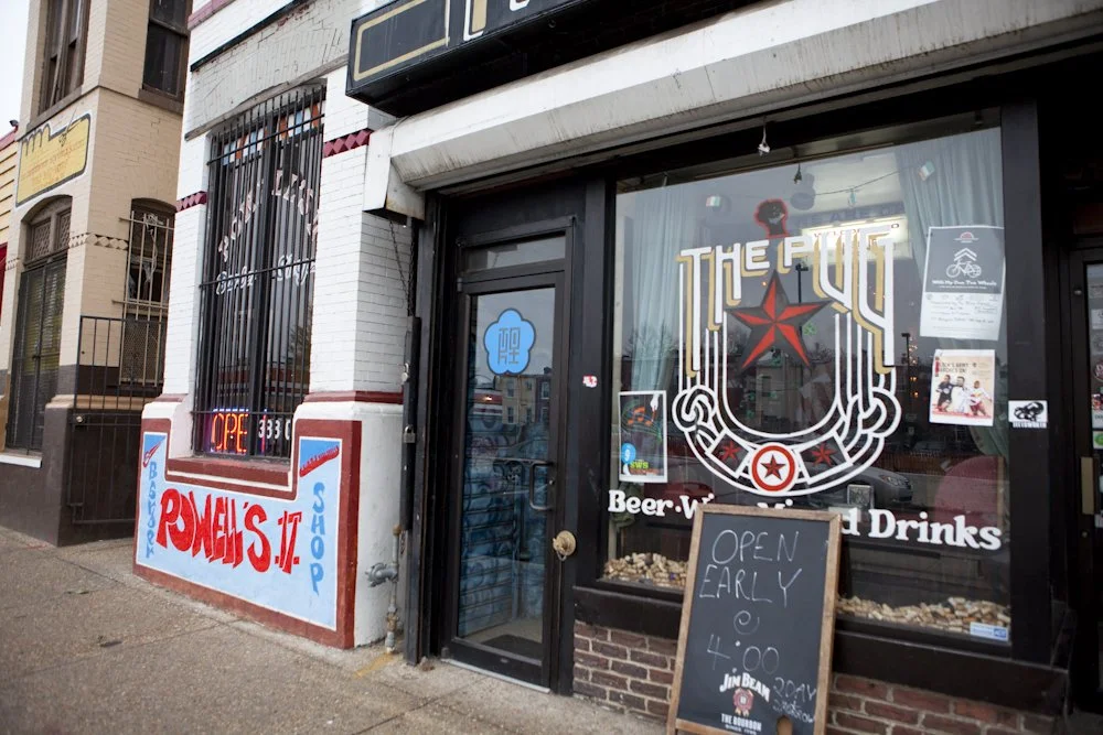 Exterior of a bar or restaurant with a large logo on the window reading 'The Fill' and a chalkboard sign outside stating 'Open early at 4:00.' The sidewalk in front has a red, blue, and white painted sign with the words 'Ronnie's' and 'Shop.'