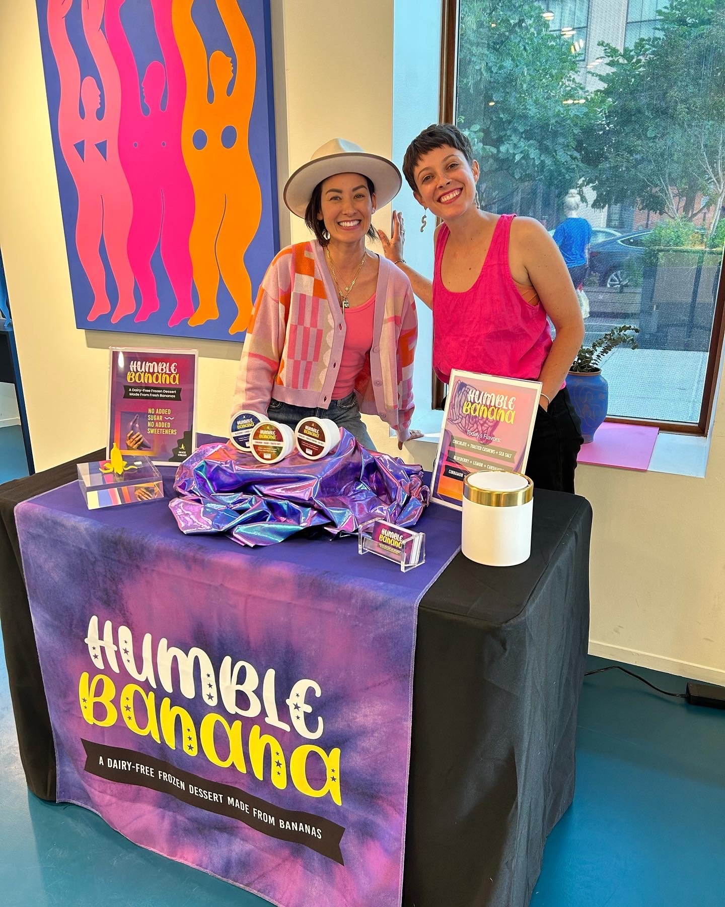 Two women smiling at a promotional table for Humble Banana, a dairy-free frozen dessert made from bananas. The table has a purple tablecloth with the product's name and description. Behind them is vibrant artwork with abstract human figures in pink and orange, set against a blue background. One woman is wearing a pink tank top and the other has a pink patterned cardigan and a white hat. There are products, signs, and a wrapped gift on the table, with a window showing outside greenery.