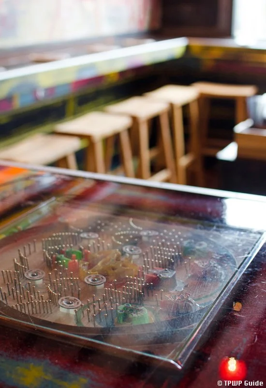 Close-up of a pinball machine with a glass cover. In the background, there are wooden bar stools along a counter in a colorful, casual restaurant or bar setting.