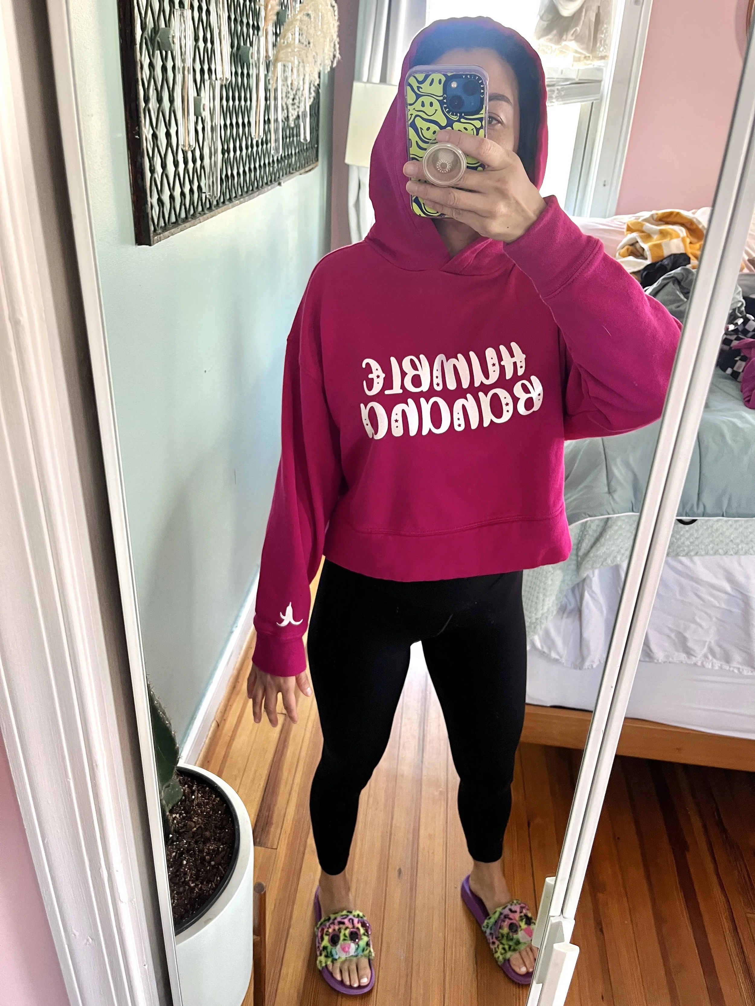 A woman taking a mirror selfie wearing a pink hoodie with the words 'Beautiful & Humble' printed upside down on it, black leggings, and colorful slippers with animal faces. The room has wooden floors, a bed with discarded clothes, a window, and a round plant pot with a cactus.