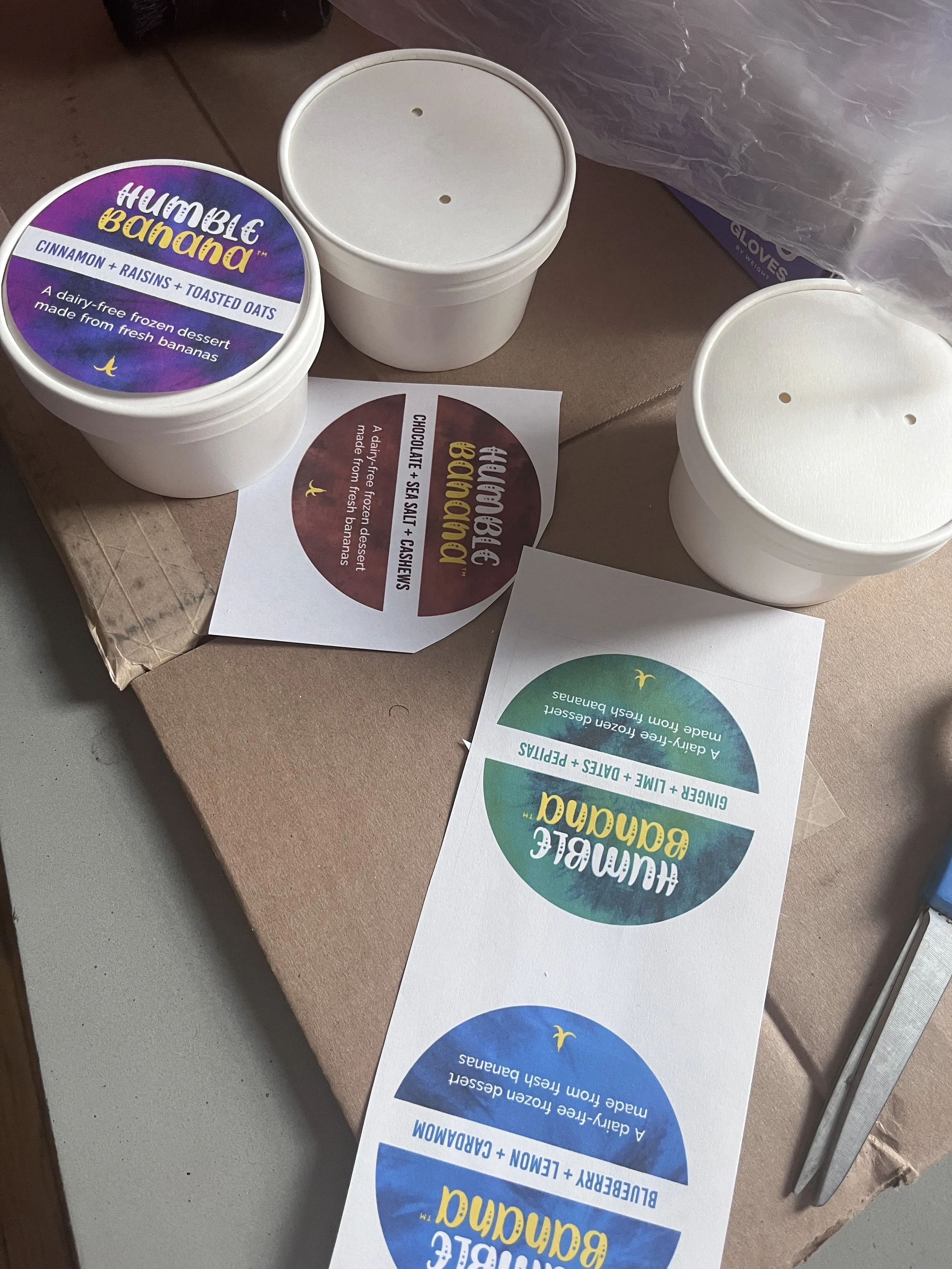 Three containers with yogurt, one labeled 'HumBlic Banana' flavor, and two plain white containers, along with colorful flyers about frozen dessert options on a brown surface.