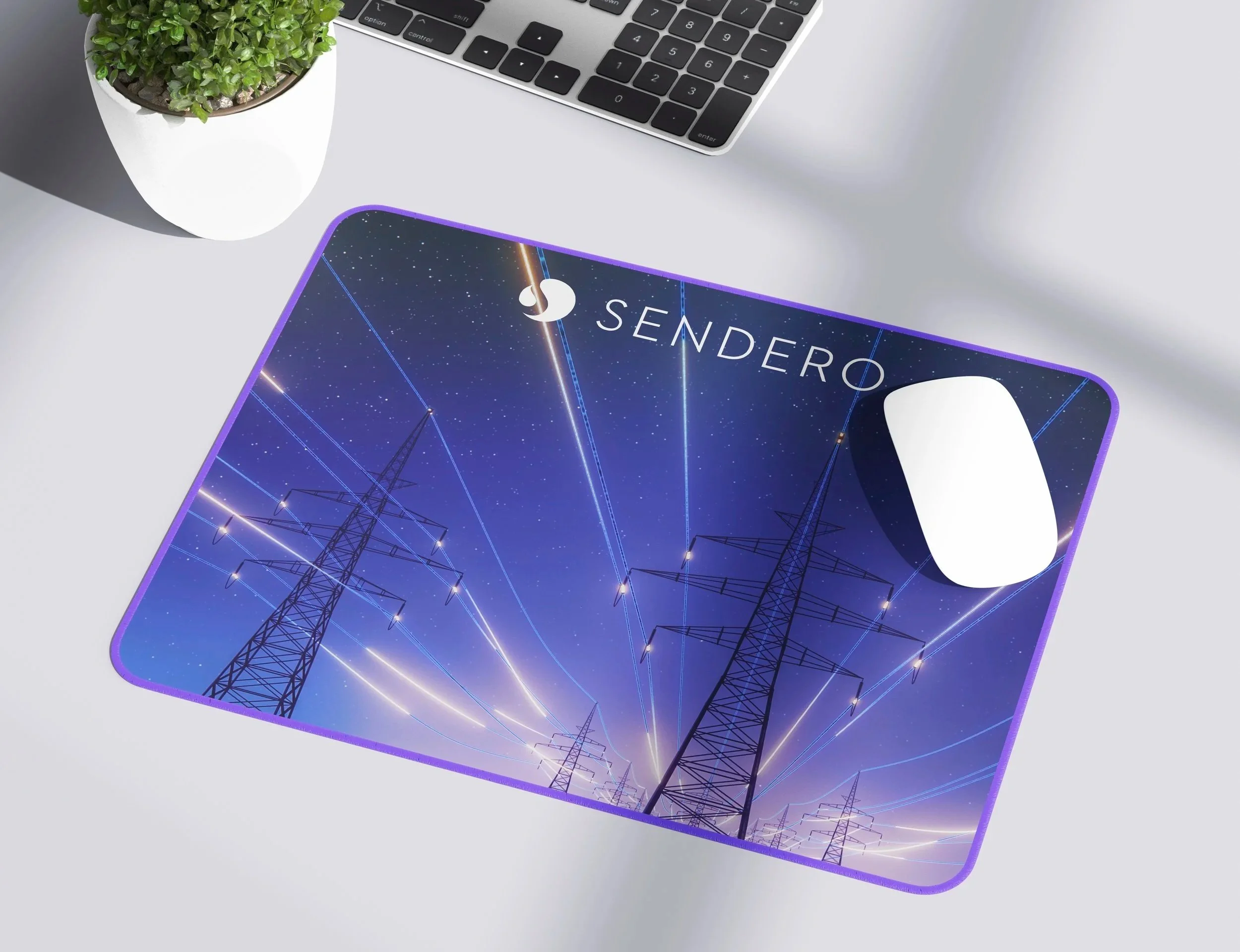 A desk scene with a laptop keyboard, a potted green plant, a wireless white mouse, and a purple mouse pad displaying an image of power lines at night with stars in the sky and the word "SENDERO".