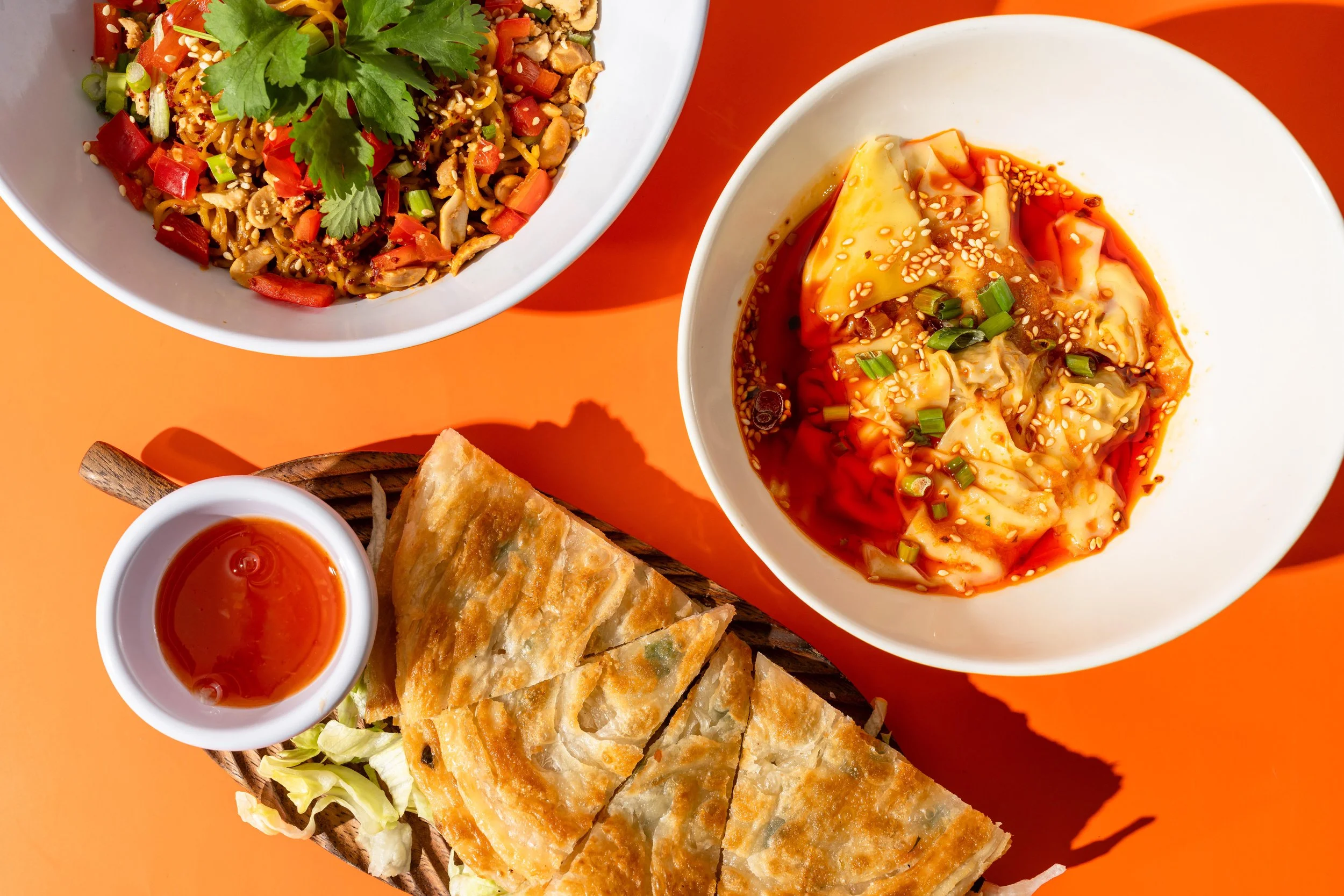 A white bowl of dumplings topped with sauce and sesame seeds, next to a basket of sliced fried bread and a small dish of dipping sauce, with a larger white bowl of noodle salad topped with cilantro and chopped vegetables on an orange background.