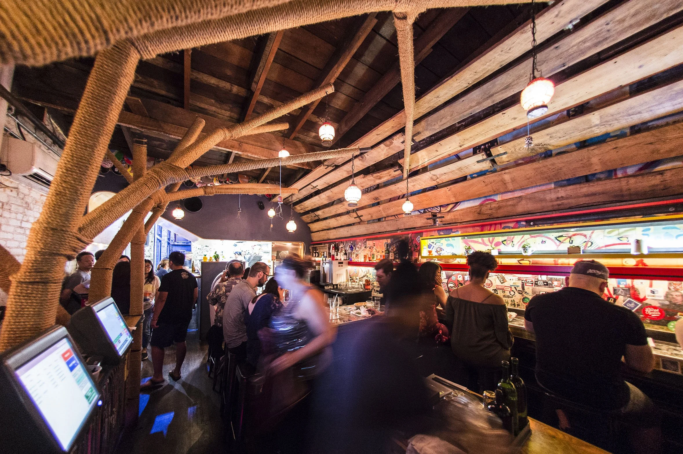 Crowded bar or restaurant with people standing and chatting, illuminated by hanging lanterns, with a wooden ceiling and a vibrant mural or artwork on the wall.