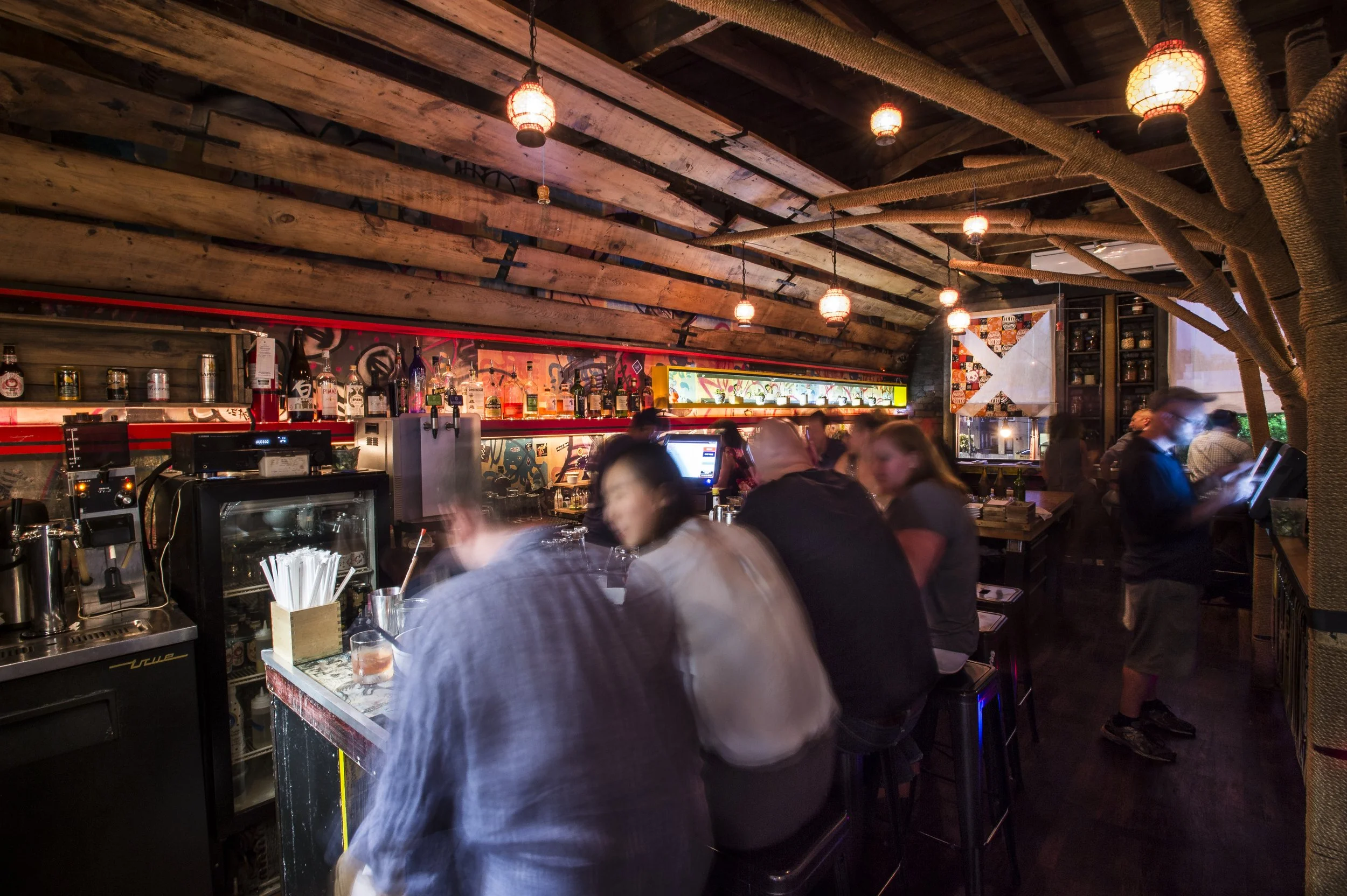 Interior of a lively bar or restaurant with wooden ceiling beams, hanging lanterns, and a decorated wall with bottles and artwork. People are socializing and using their phones.