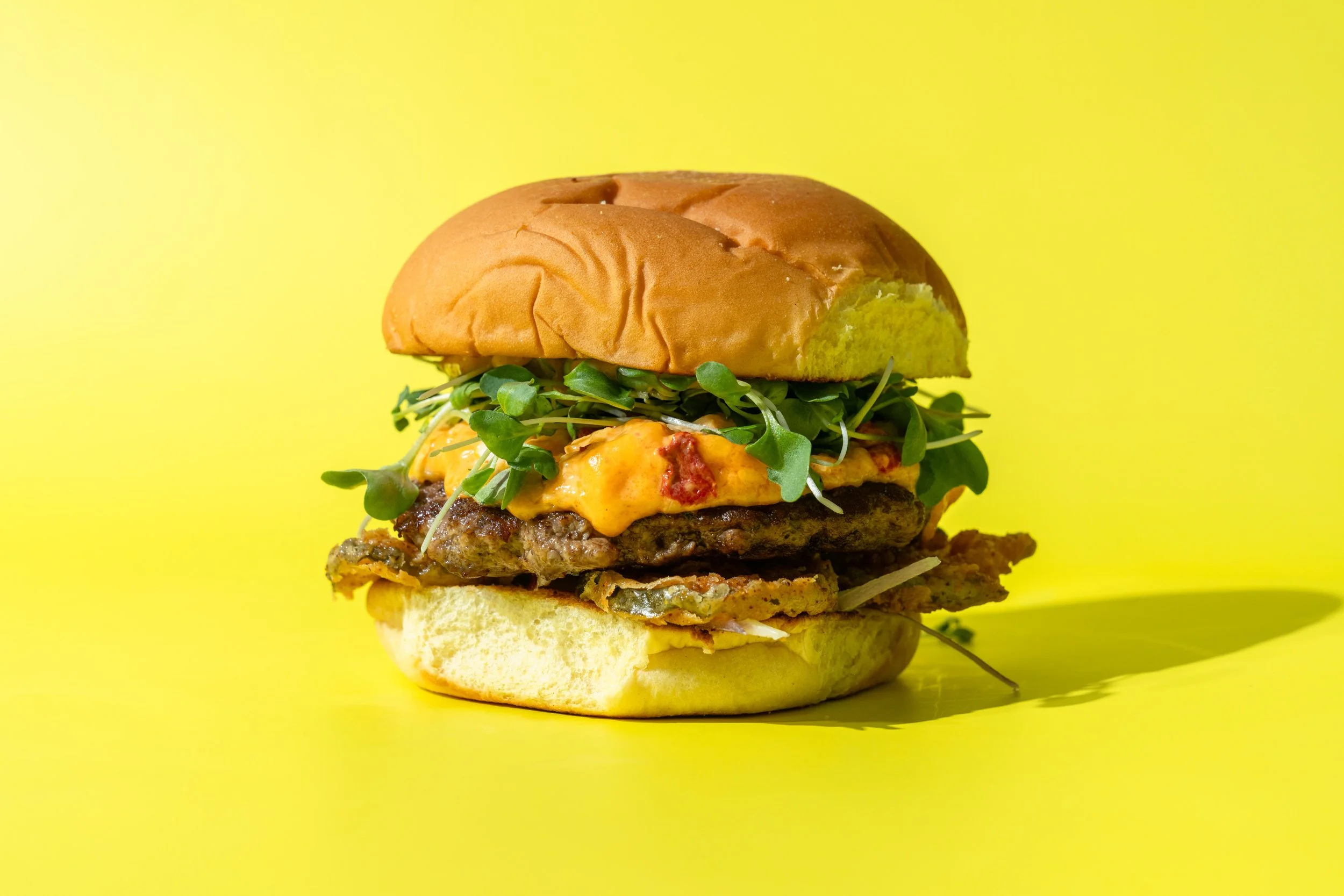 A close-up of a hamburger with a bun, leafy greens, melted cheese, a beef patty, and condiments, against a bright yellow background.