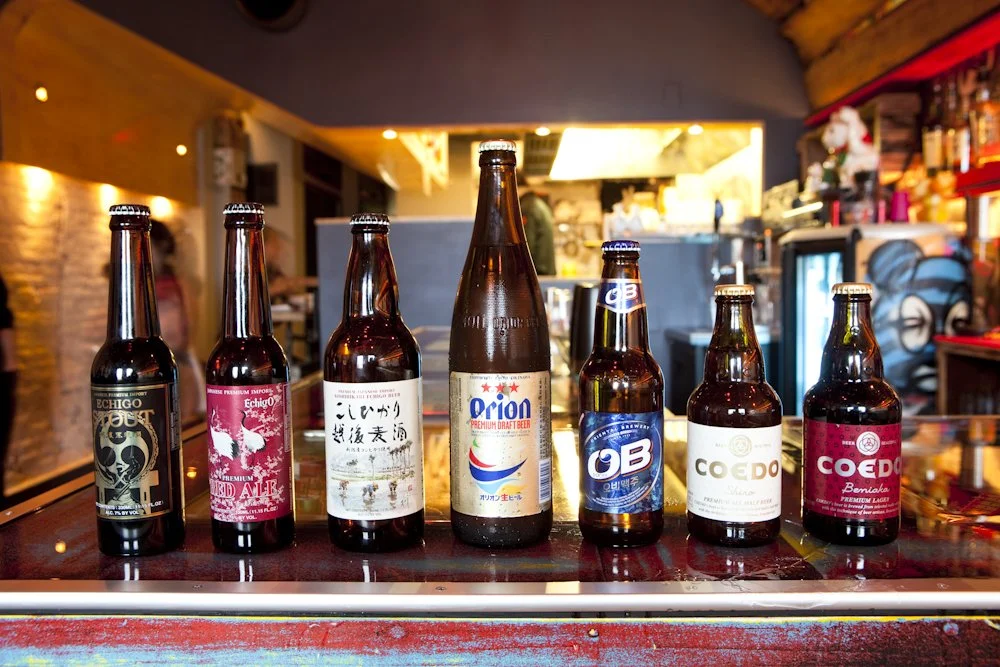 Six different bottles of beer lined up on a bar counter inside a lively bar or pub with colorful interior decor.