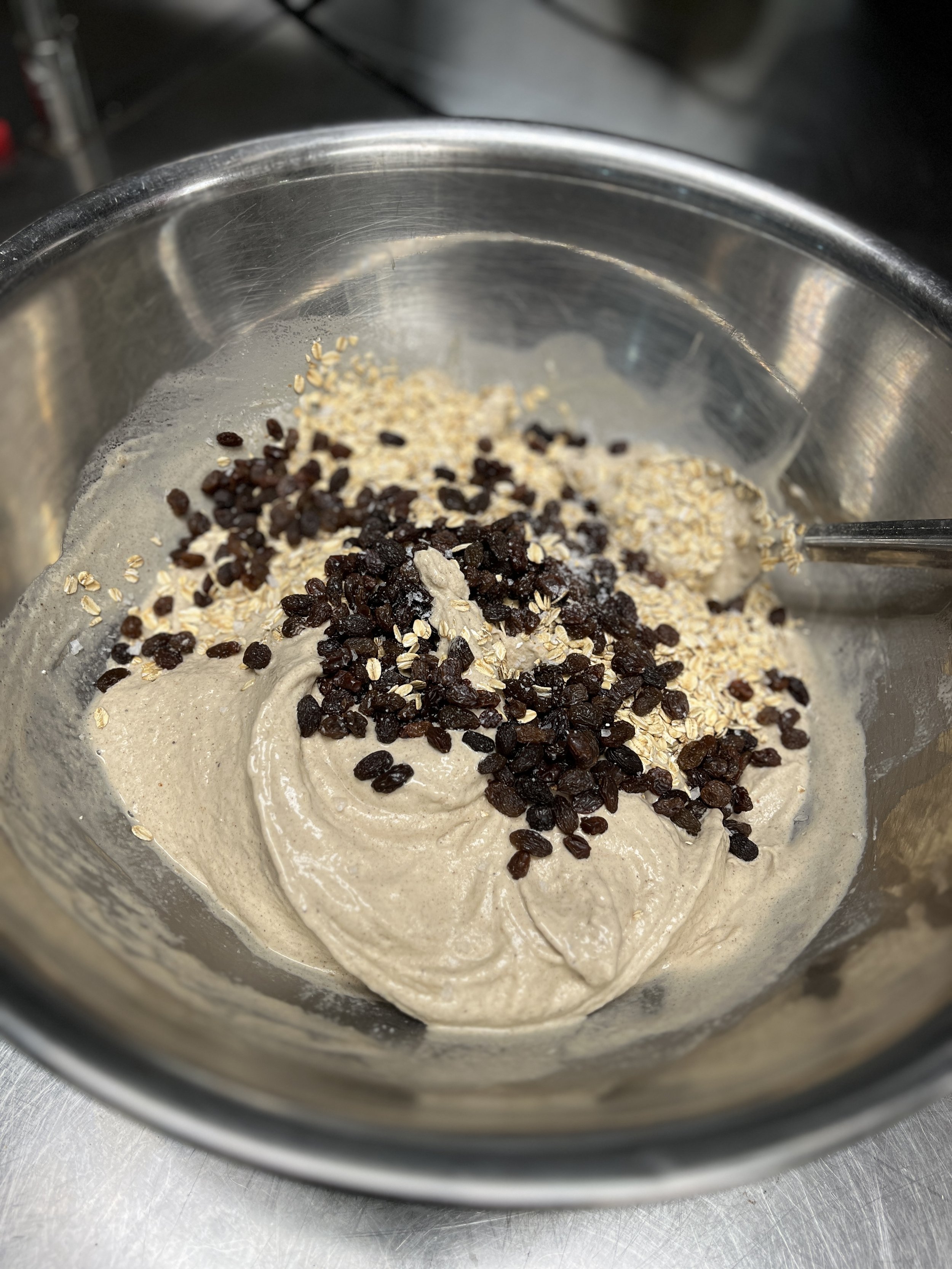 A mixing bowl containing a batter with oats and raisins on top, with a spoon partially submerged, placed on a metallic surface.