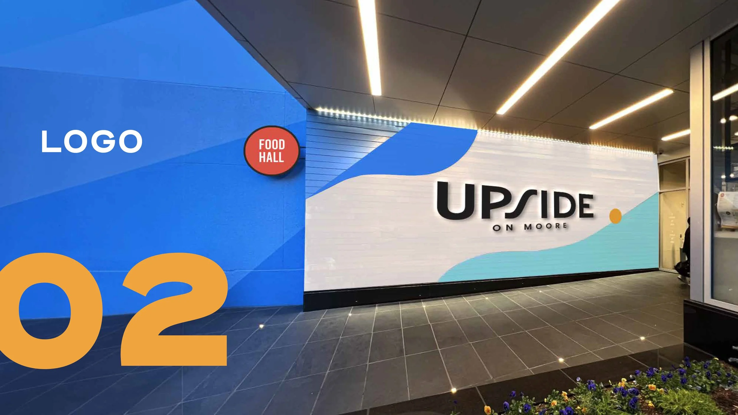 Modern building interior with a large blue and white mural displaying the words 'UPSIDE ON MOORE' with geometric graphics, a lit ceiling, and floral decoration in the foreground.